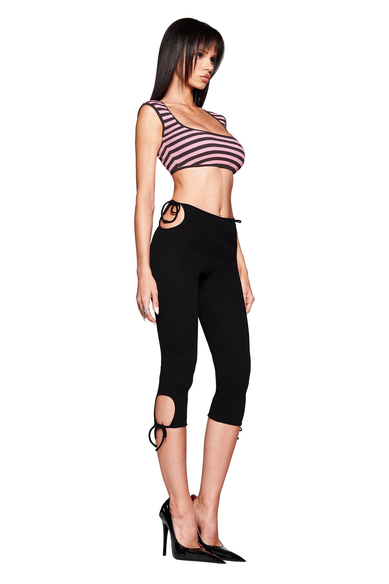 A woman in profile wears a pink and black striped crop top, I.AM.GIA YUNA CAPRI - BLACK body-con mid rise leggings with flower cut out details, and black high-heeled shoes, set against a white background.