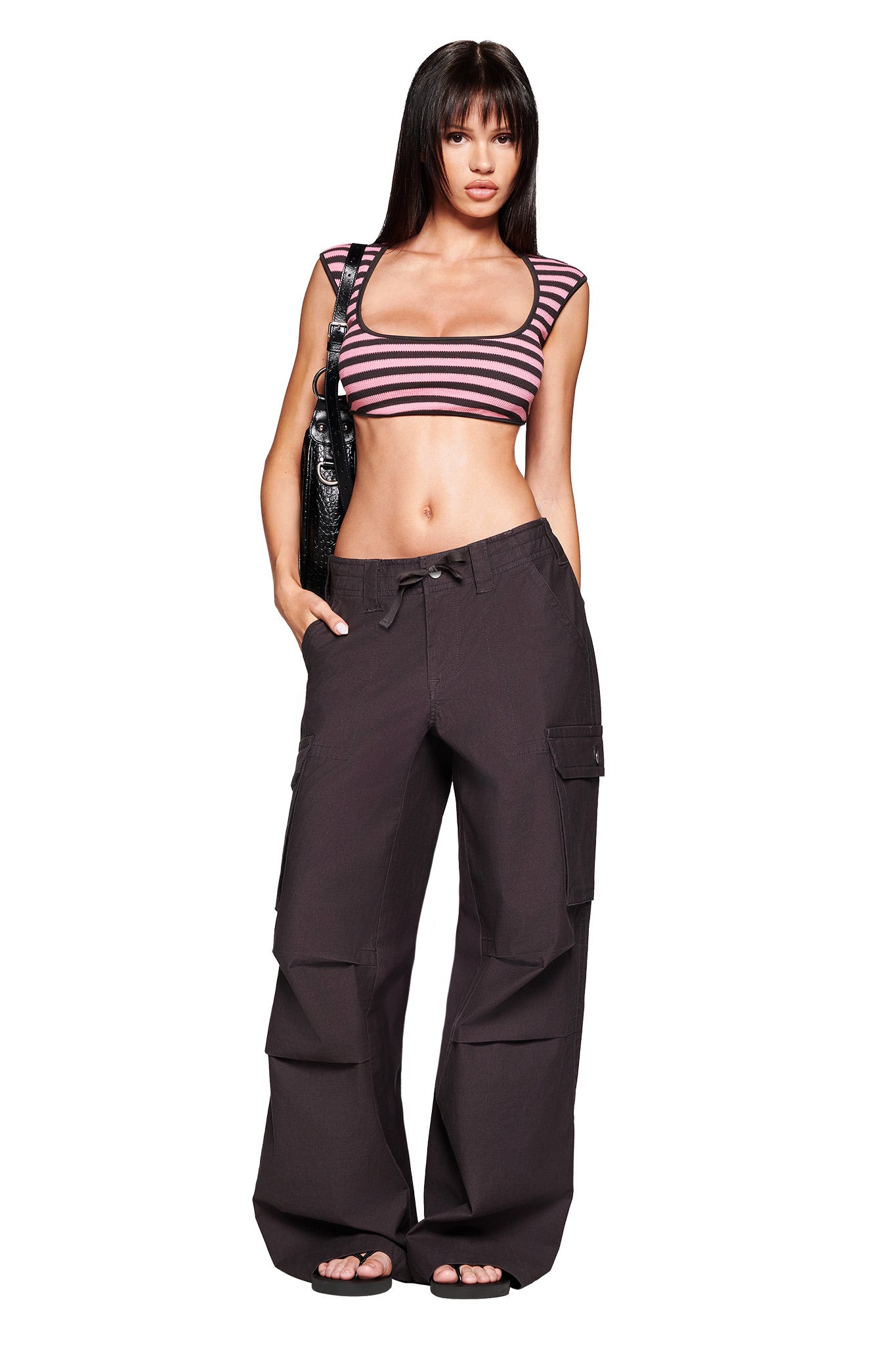 A woman stands confidently in the I.AM.GIA RICA TOP - PINK/GREY, paired with loose black cargo pants and sandals. She has long dark hair, carries a black shoulder bag, and poses against a plain white background.