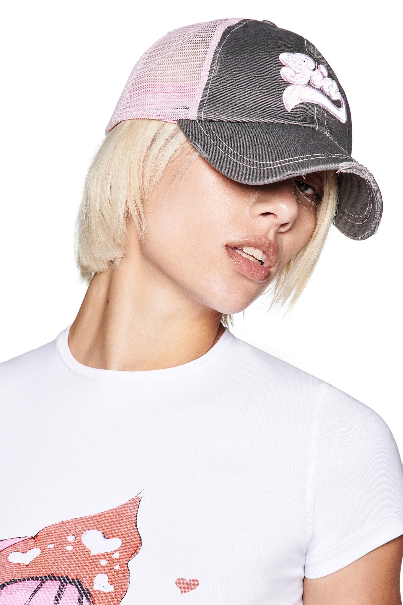 A person with short blonde hair wears the I.AM.GIA SHILOH HAT in grey and baby pink, featuring a distressed trucker style and custom logo, paired with a white tee with a pink graphic, looking slightly to the side with head tilted down.