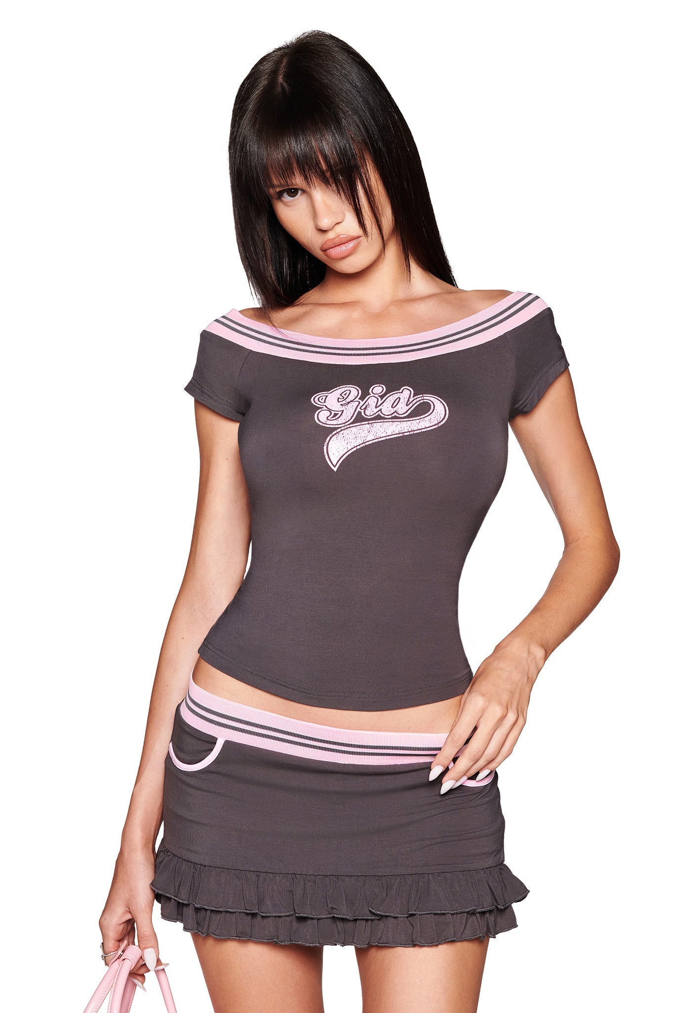 A woman with straight dark hair wears the I.AM.GIA ELLIS TOP in grey bamboo jersey, showcasing a pink collegiate logo, paired with a matching ruffled mini skirt. She holds a light pink accessory against a white background.