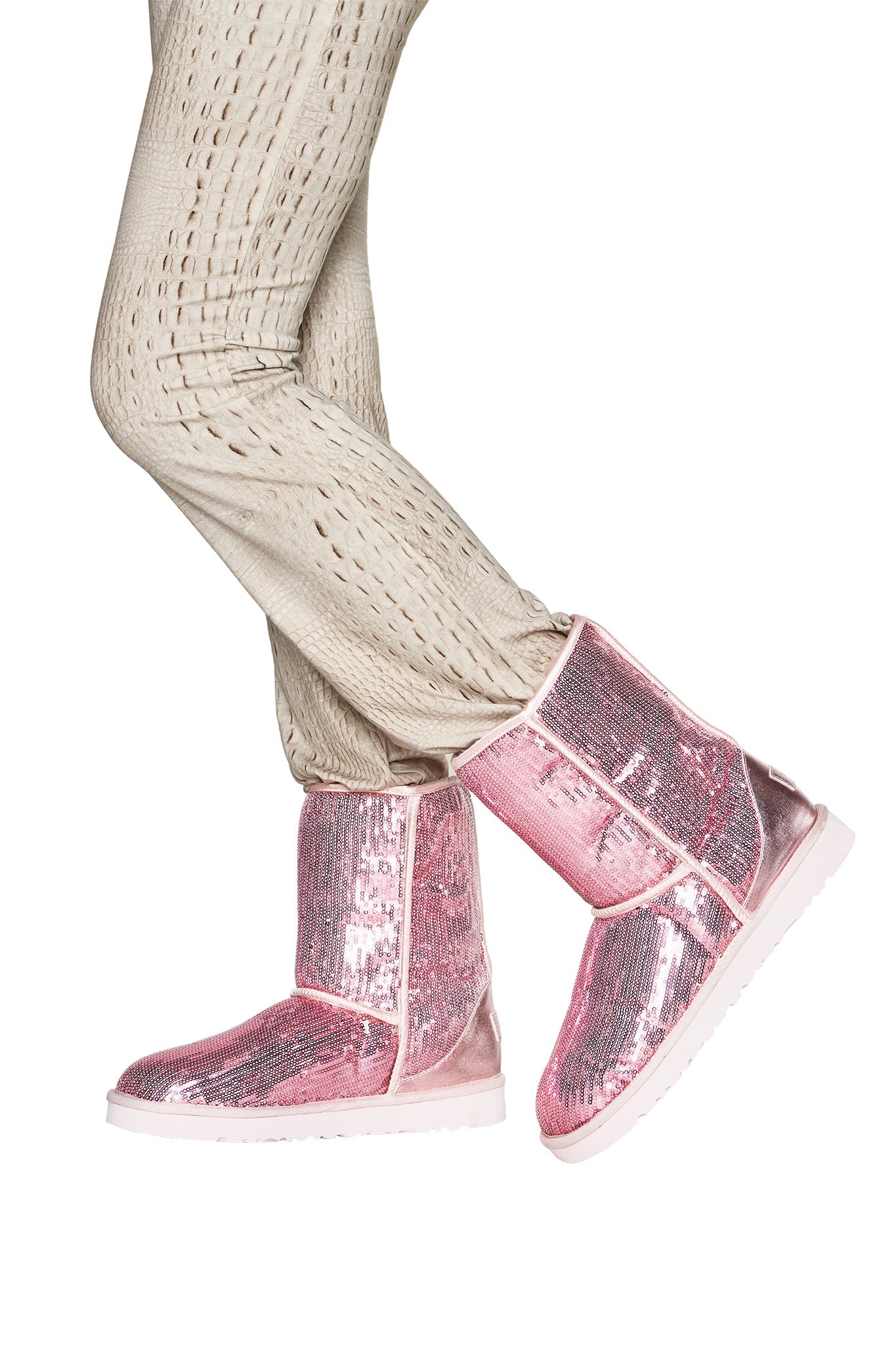 Wearing the I.AM.GIA ELVIRA SEQUIN BOOT in pink, featuring a shiny sequin finish and soft faux fur lining, a person stands against a white background with one leg crossed in front of the other, paired with beige textured pants.