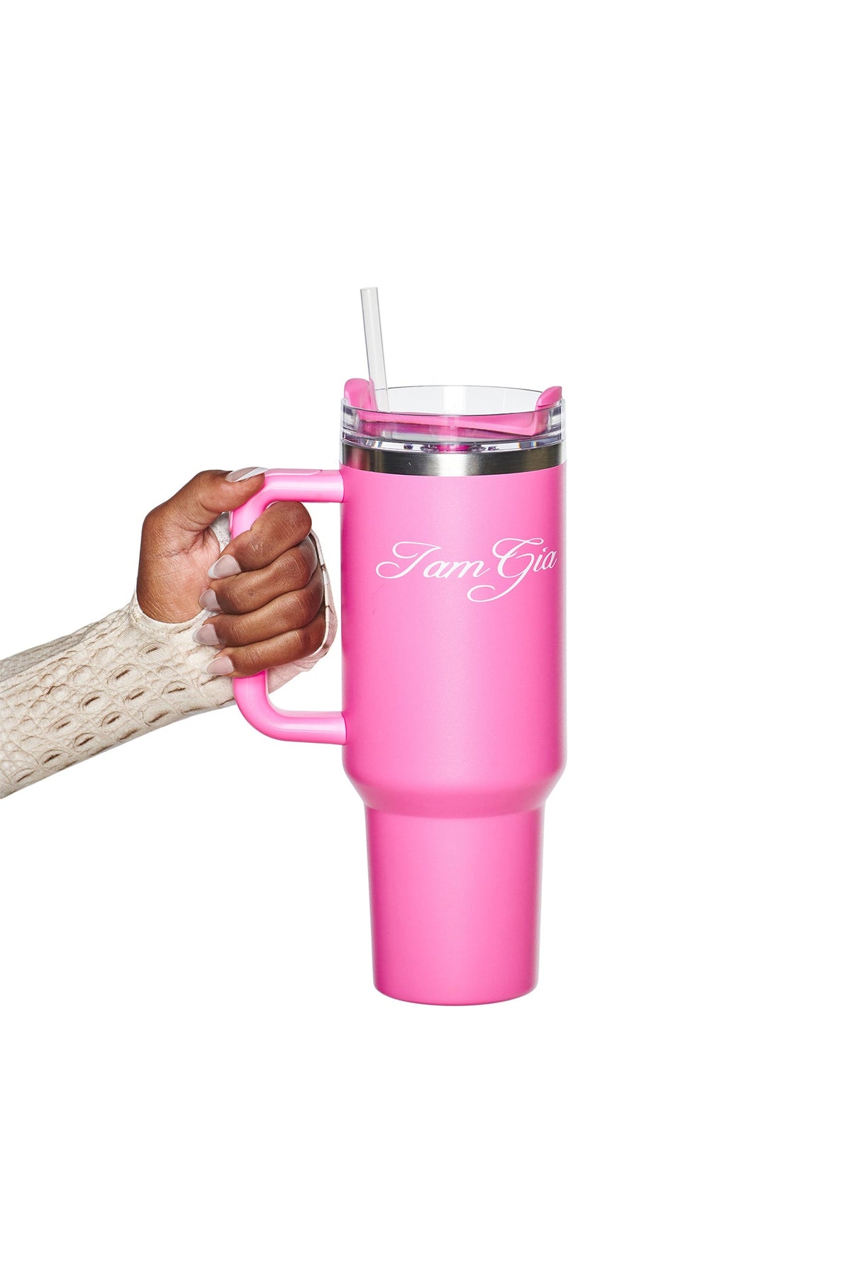 A hand holds the BLARE CUP - PINK, an insulated stainless steel tumbler from I.AM.GIA, featuring a clear lid, straw, and the brand name “I.AM.GIA” in white script on the side. The person’s beige textured sleeve is visible in the shot.