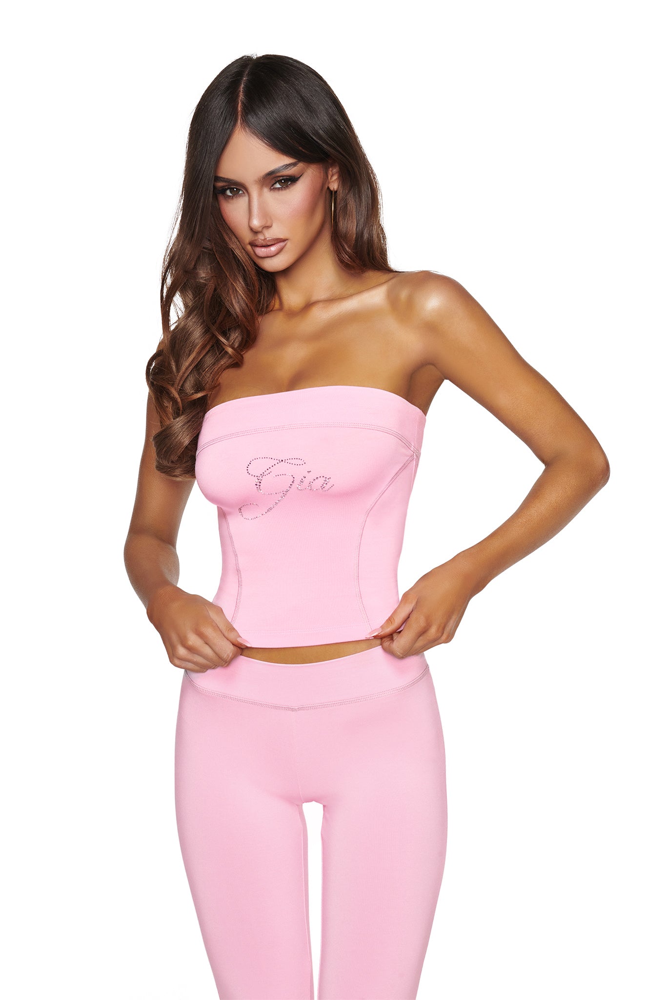 A woman with long brown hair wears the I.AM.GIA BLARE DIAMONTE BOOB TUBE in baby pink, featuring rhinestone text, along with matching pink leggings, standing against a plain white background.