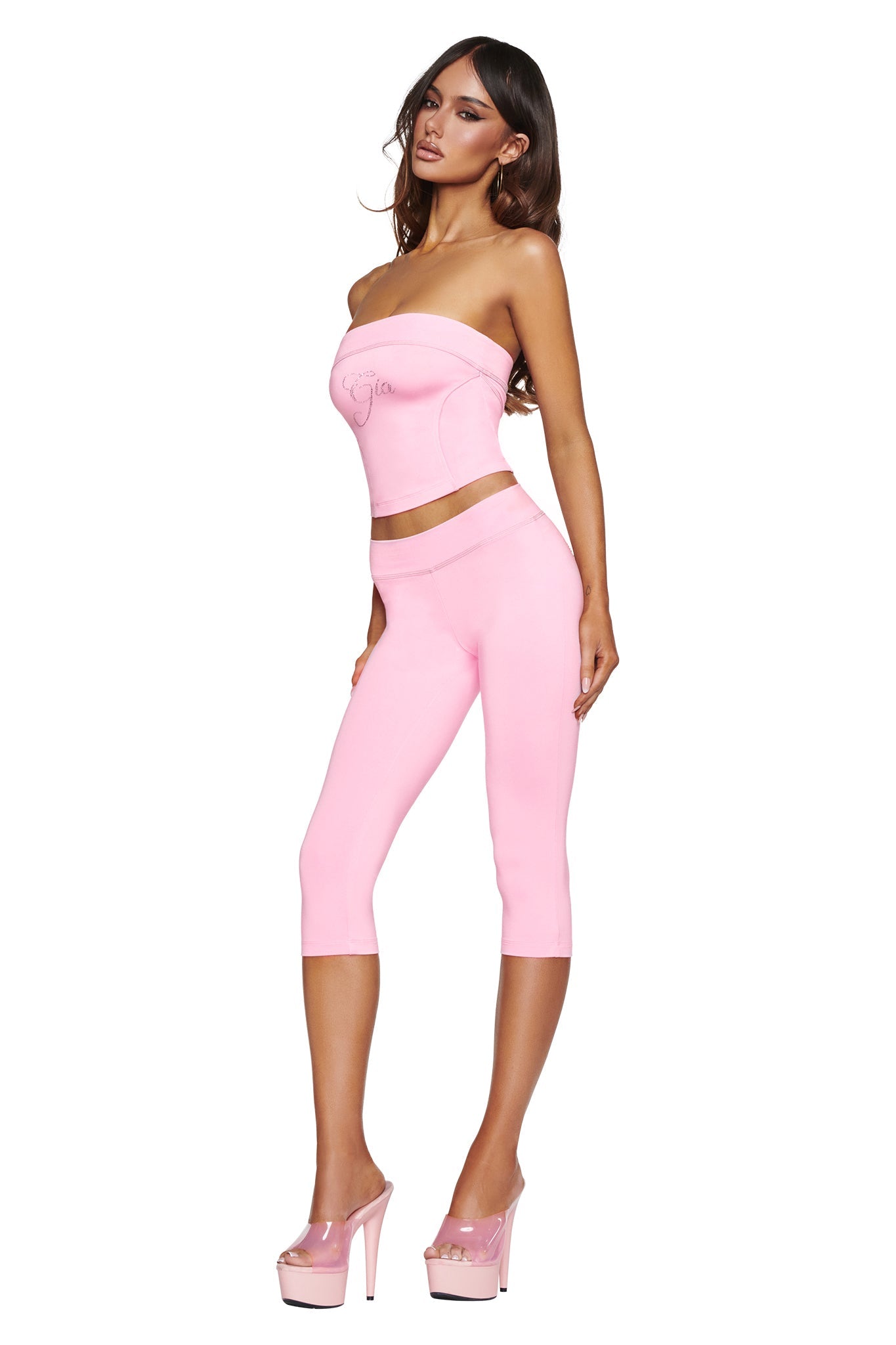 A woman poses confidently against a white background wearing a strapless pink top, I.AM.GIA’s BLARE LOW RISE DIAMONTE CAPRI in baby pink, and matching pink platform heels.