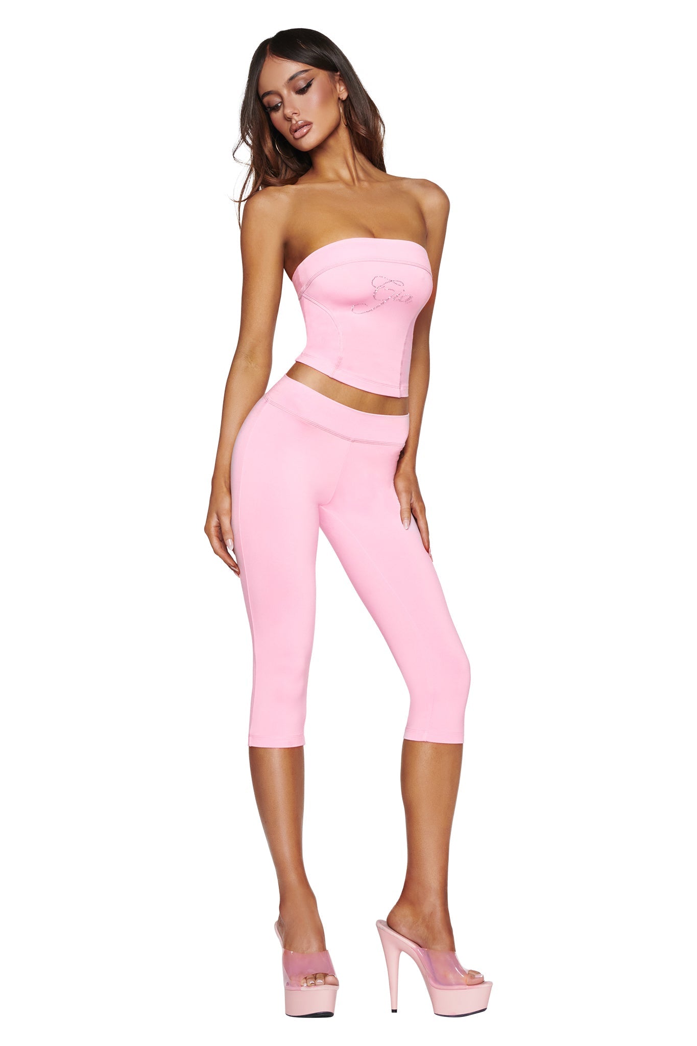 A woman with long brown hair stands against a white background, wearing the BLARE DIAMONTE BOOB TUBE in baby pink by I.AM.GIA and matching pink capri leggings, paired with high platform heels.