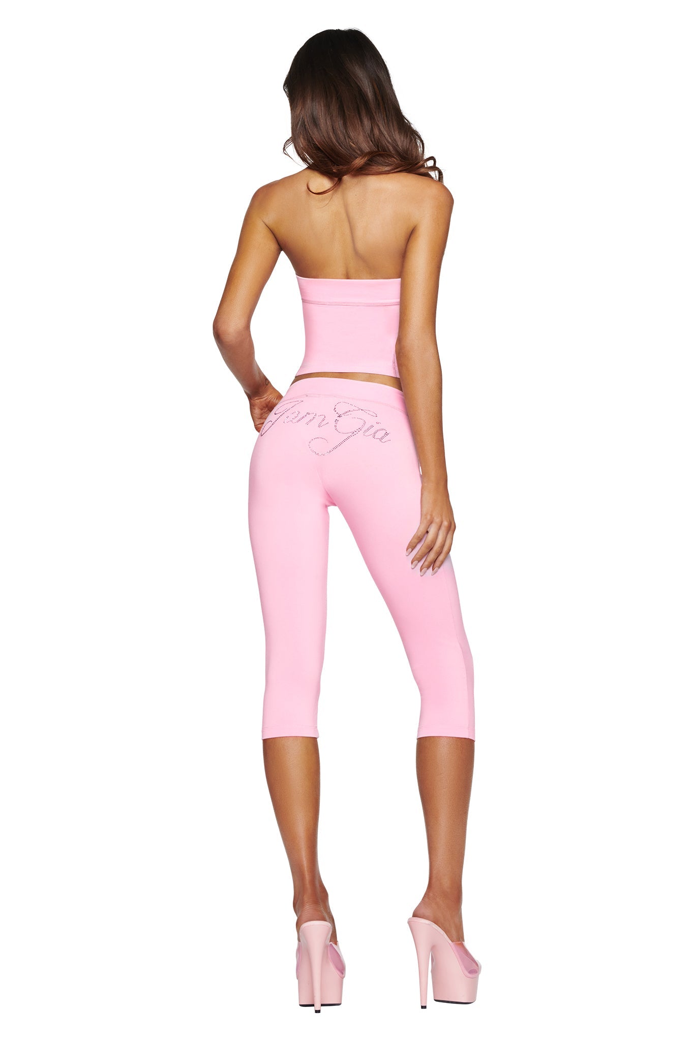 A woman stands with her back to the camera, wearing the I.AM.GIA BLARE DIAMONTE BOOB TUBE in baby pink, along with matching pink capri leggings featuring script text on the back and high pink platform heels, against a white background.