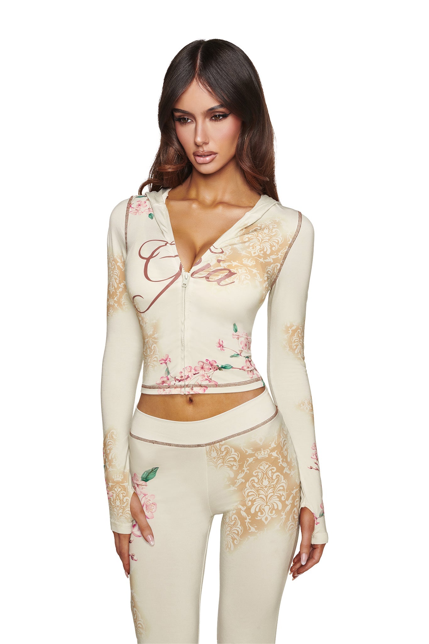 A woman wearing the I.AM.GIA BLARE HOODIE - BLOSSOM, a fitted cream-colored two-piece set with a zip-up bamboo hoodie and matching pants adorned with gold patterns and pink floral designs, standing against a white background.