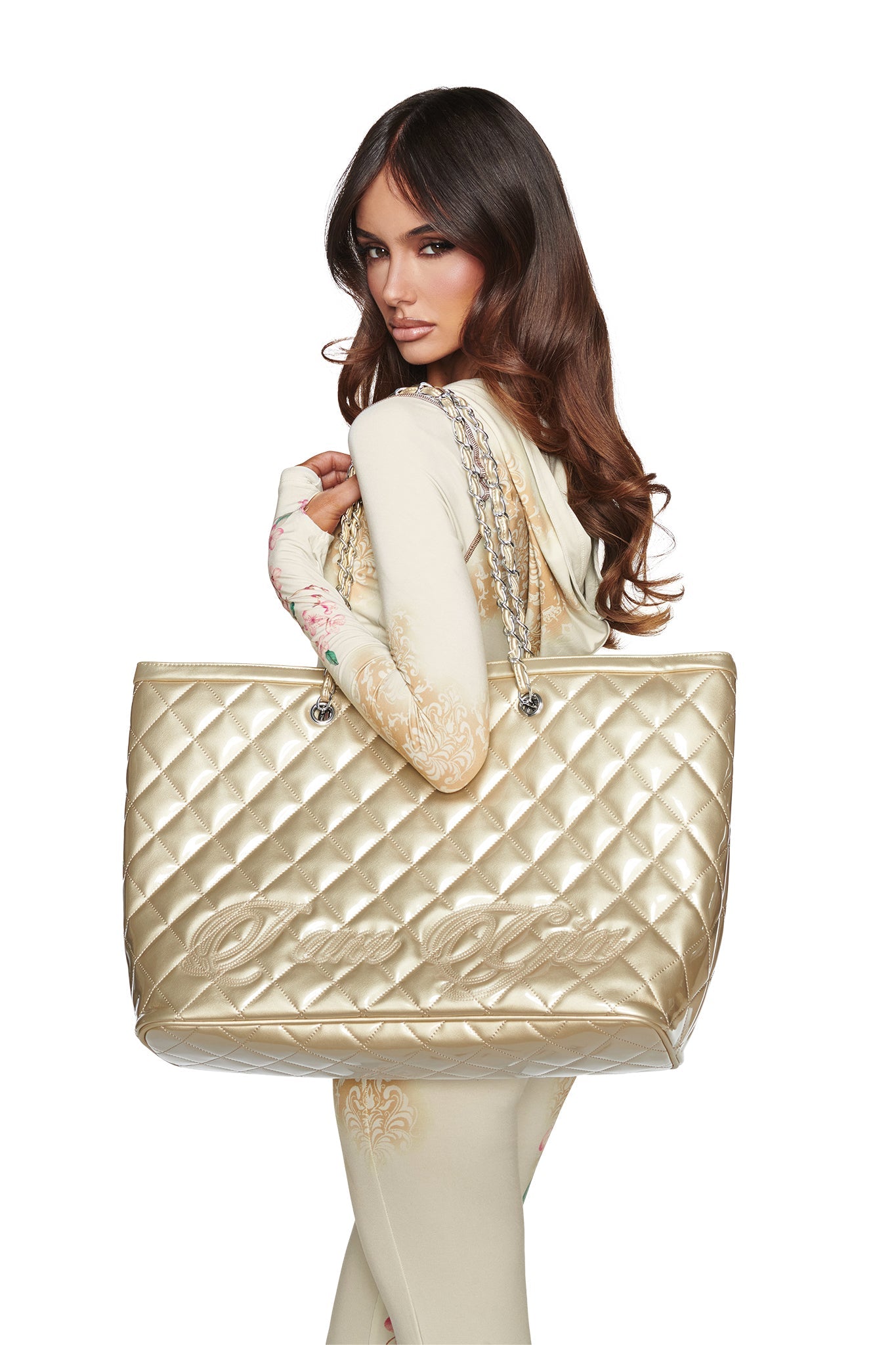 A woman with long brown hair in a cream outfit carries the I.AM.GIA DANNI TOTE BAG in gold, featuring oversized metallic design and chain link straps, posing against a white background.
