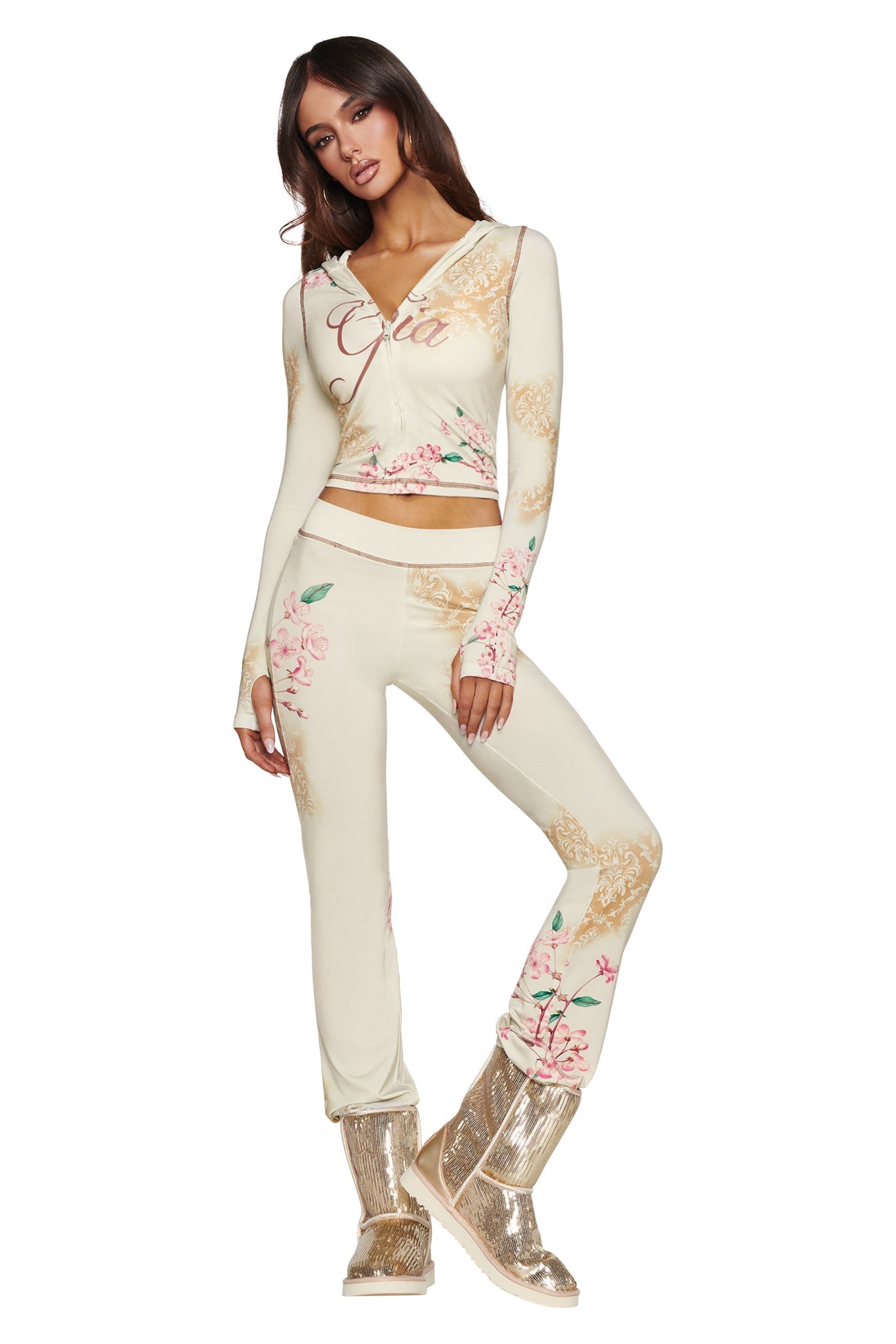A woman poses with one hand on her hip in a cream-colored floral lounge set featuring long sleeves and pants. She completes the look with the I.AM.GIA ELVIRA SEQUIN BOOT in gold, which sparkles with shimmering sequins and features a cozy faux fur lining. The background is plain white.