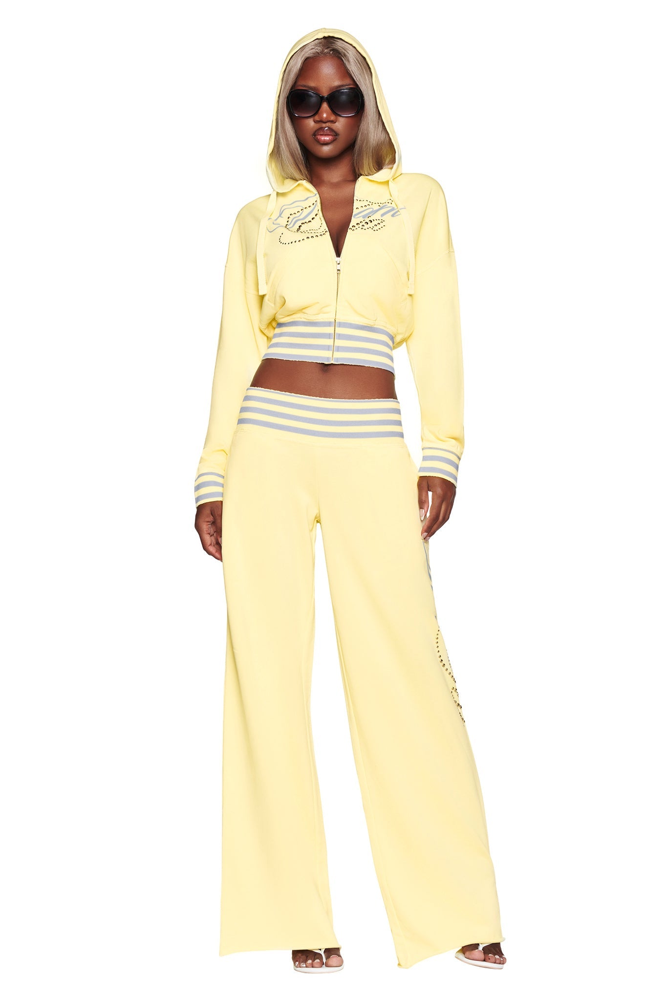 A woman wearing the I.AM.GIA VEDA PANT in yellow, paired with a matching zip-up hoodie, sunglasses, and sandals, stands against a white background.