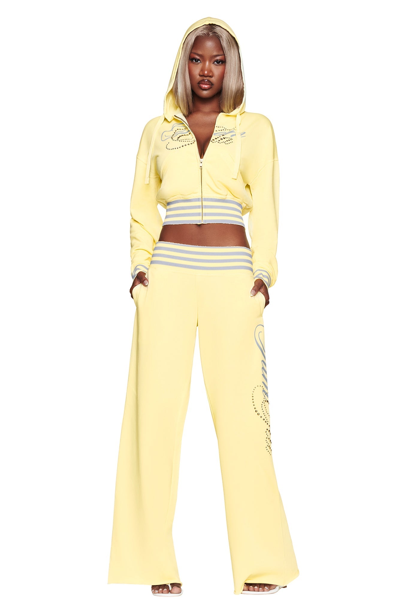 A woman wearing the I.AM.GIA VEDA PANT in yellow, featuring a low-rise, wide-leg silhouette and gray and white striped waistband, stands with her hands in her pockets against a white background.