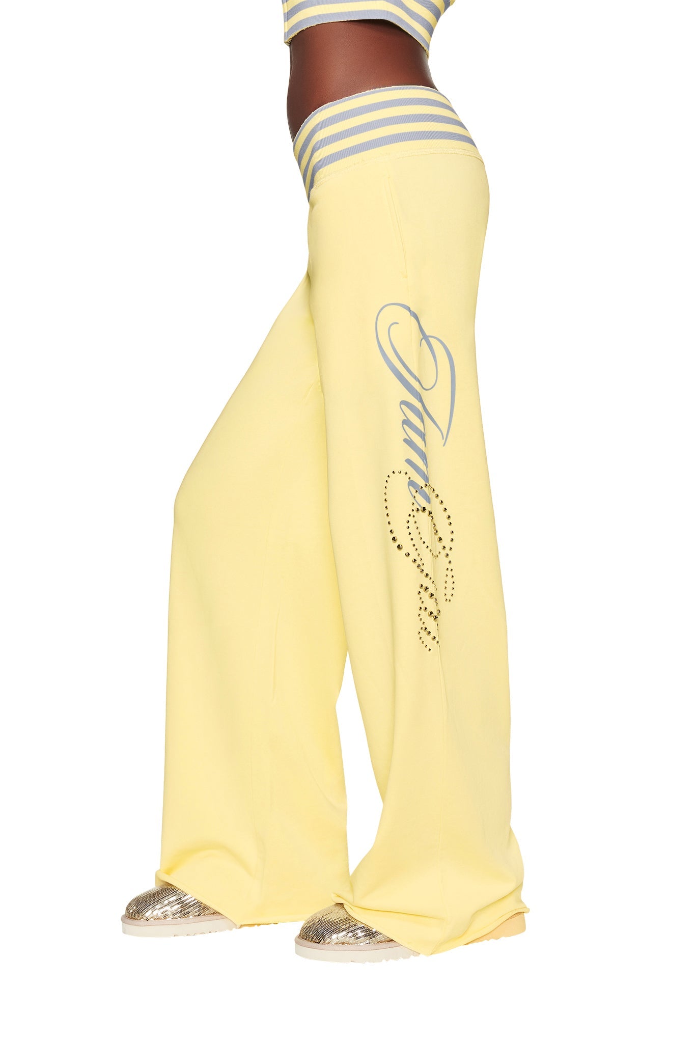 A person wearing the I.AM.GIA VEDA PANT in yellow—low-rise, wide-leg sweatpants with “Juicy” in silver and rhinestones on the leg—paired with a striped crop top and silver glitter sneakers.