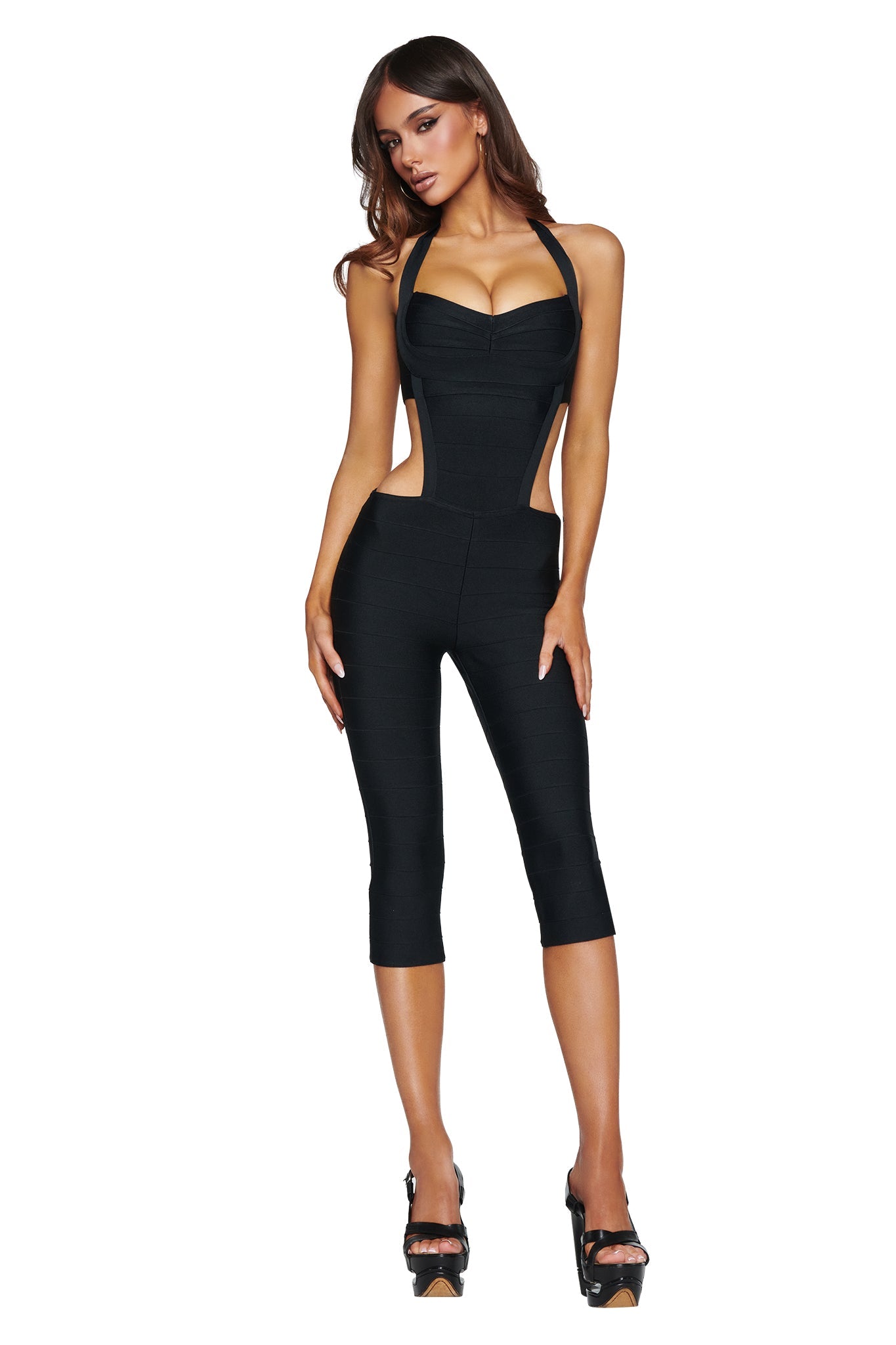 A woman poses in the I.AM.GIA EVA BANDAGE CAPRI JUMPSUIT in black, featuring side cutouts, a sleek halter neck design, and cropped legs, paired with black high heels against a white background.