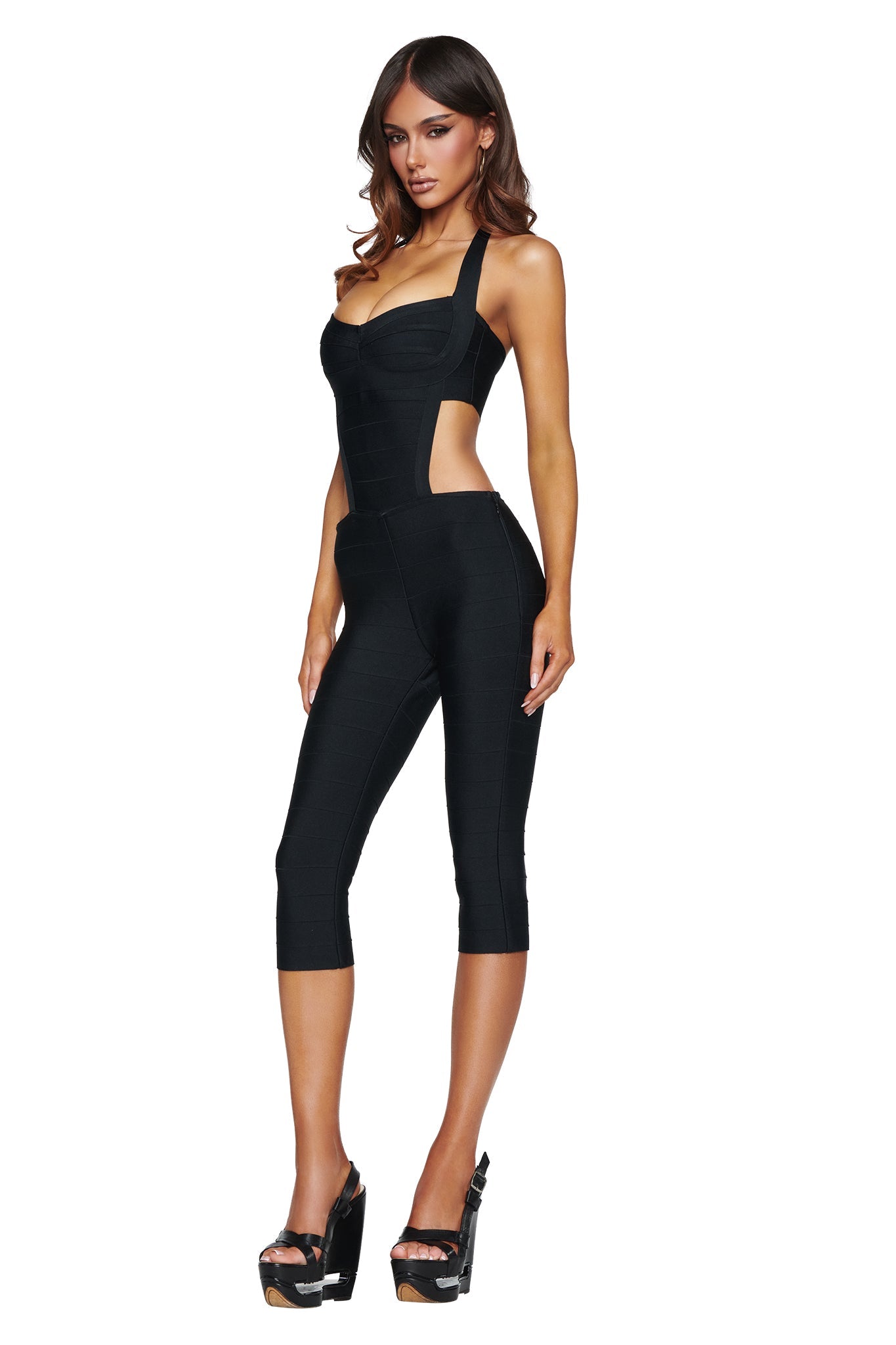Model wears the EVA BANDAGE CAPRI JUMPSUIT in black by I.AM.GIA, featuring a halter neck and side cutouts, paired with black platform heels against a white background.