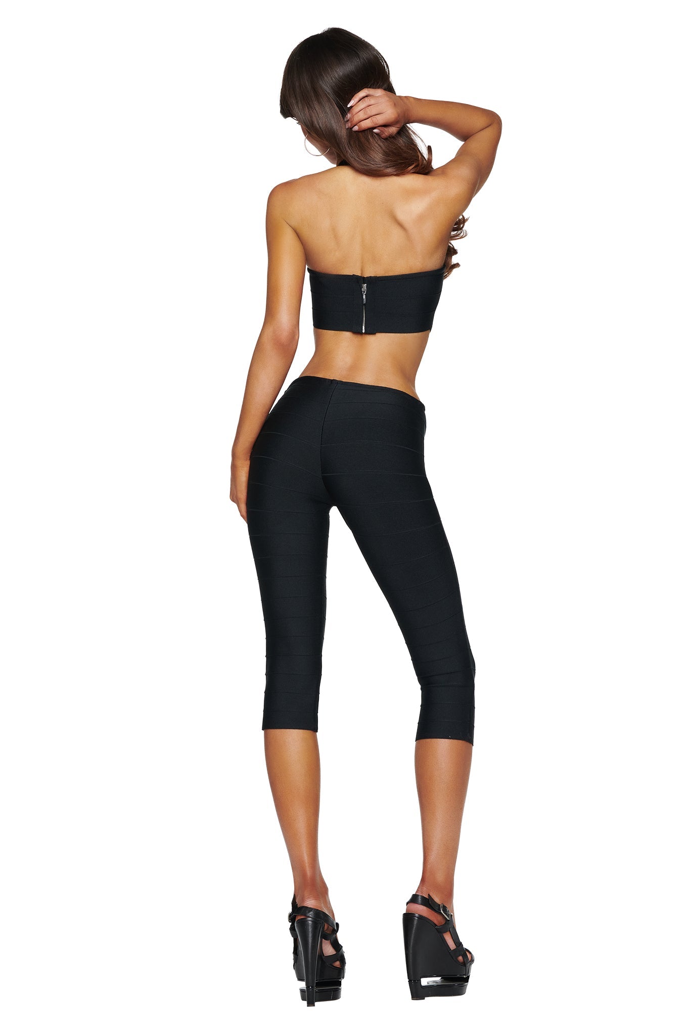 A woman stands with her back to the camera, one hand touching her hair, wearing the I.AM.GIA EVA BANDAGE CAPRI JUMPSUIT in black paired with high-heeled sandals—her sleek silhouette exuding chic sophistication.