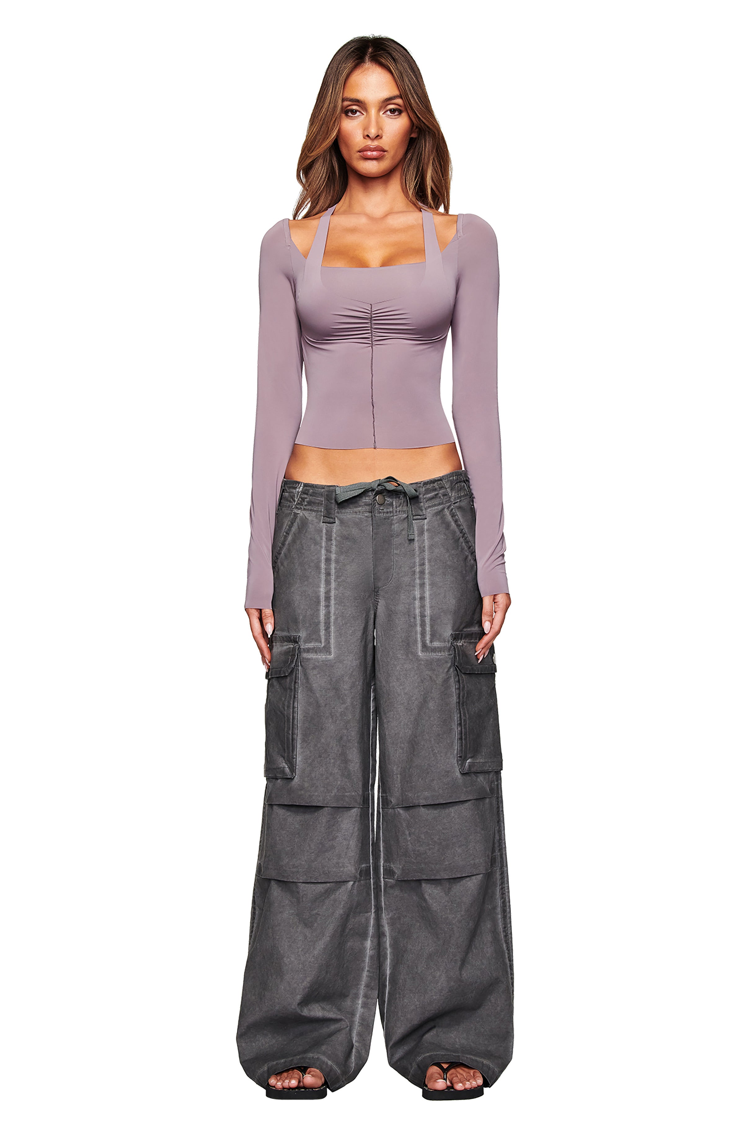 A woman faces forward in a mauve square-neck top and I.AM.GIA’s JAXXON CARGO PANT in washed black, featuring oversized pockets and a low-rise waist. She has loose, wavy hair and wears sandals against a white background.