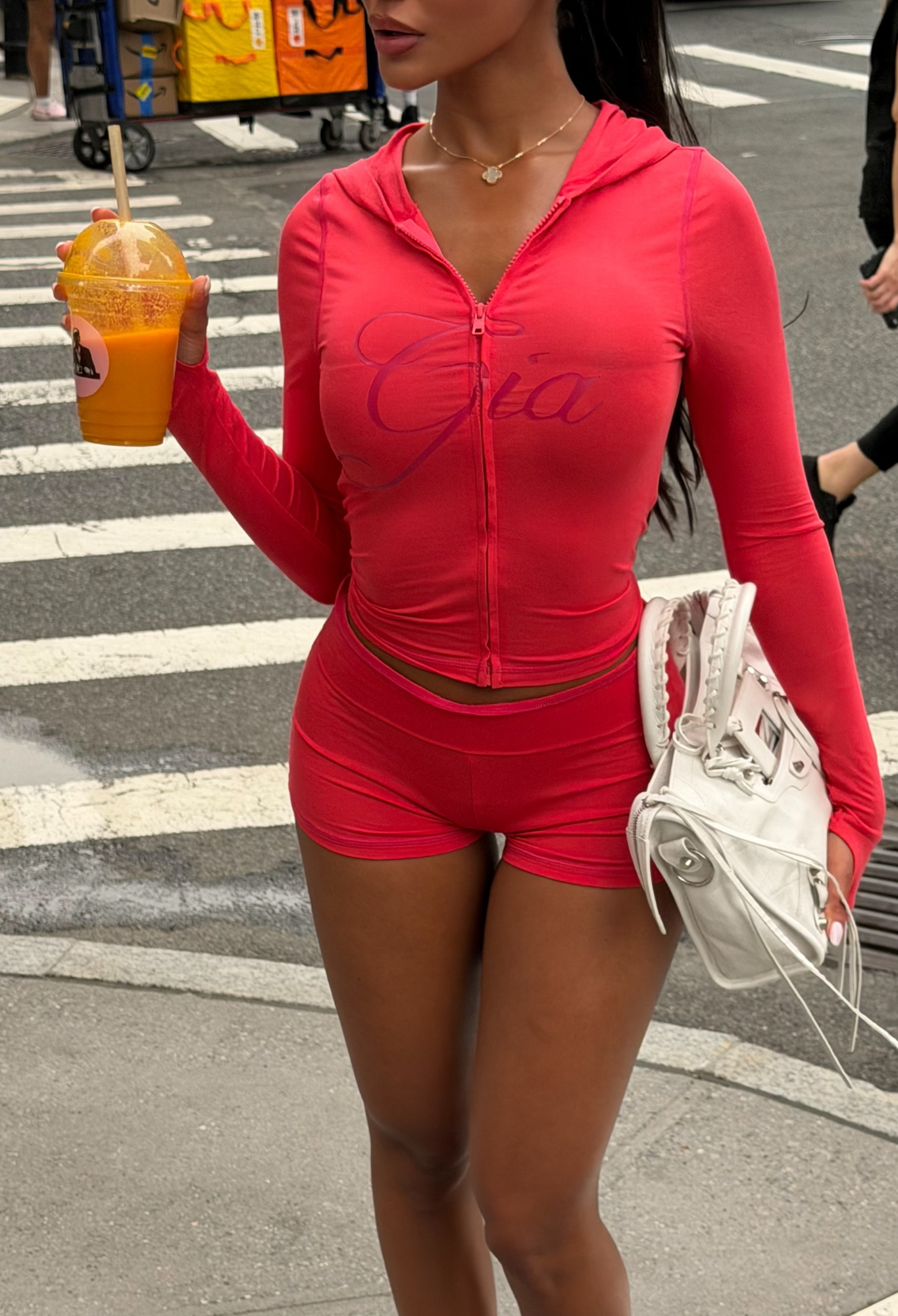 A woman in a bright pink zip-up top and the I.AM.GIA BLARE SHORT - PAPAYA, featuring an elastic waistband, holds an orange drink and a white handbag while standing on a city sidewalk near a crosswalk.