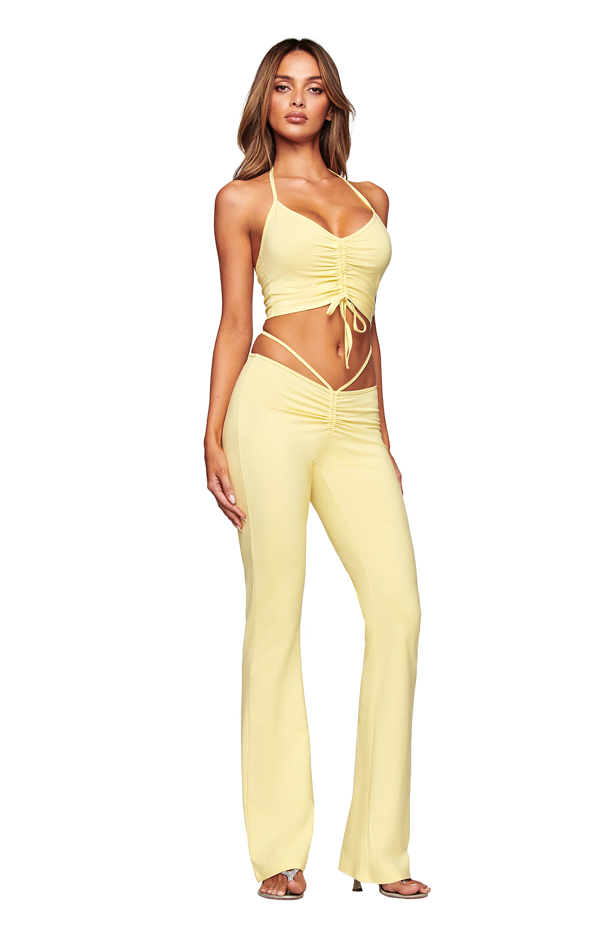 A woman stands confidently in heels against a white background, wearing the I.AM.GIA HALO TOP in butter yellow with ruching, paired with matching high-waisted flare pants.