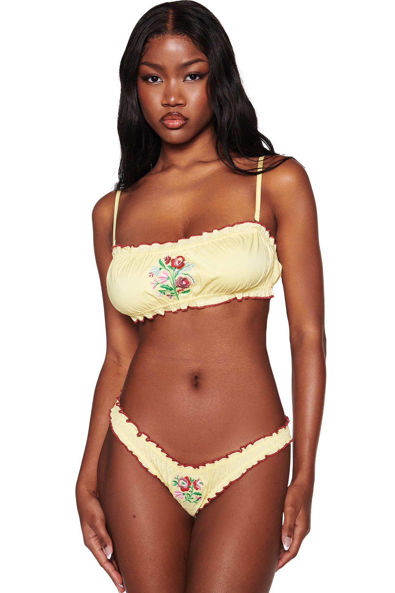The woman wears the CLOVER SET - YELLOW by I.AM.GIA, featuring exquisite Hungarian embroidery and a red trim. The bandeau top has detachable straps, and the hipster bottom complements her wavy black hair against a crisp white background.