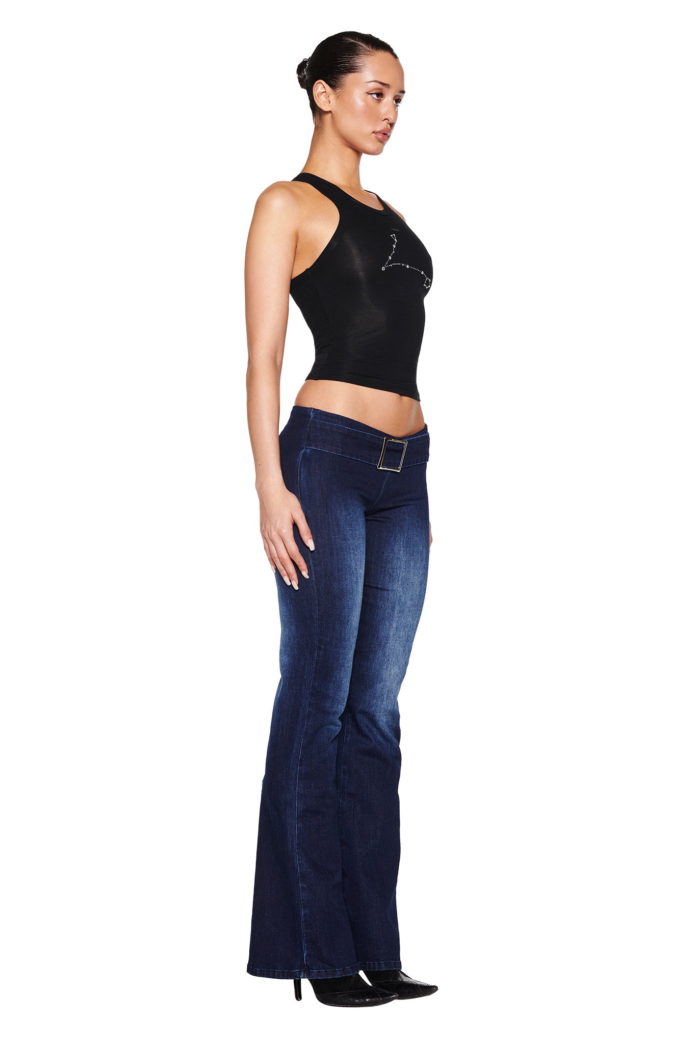 A person stands in profile on a white background, wearing a black sleeveless top with I.AM.GIA's SHARAH DENIM PANT - BLUE in dark blue stretch denim, complemented by black shoes and their hair slicked back into a sleek bun.