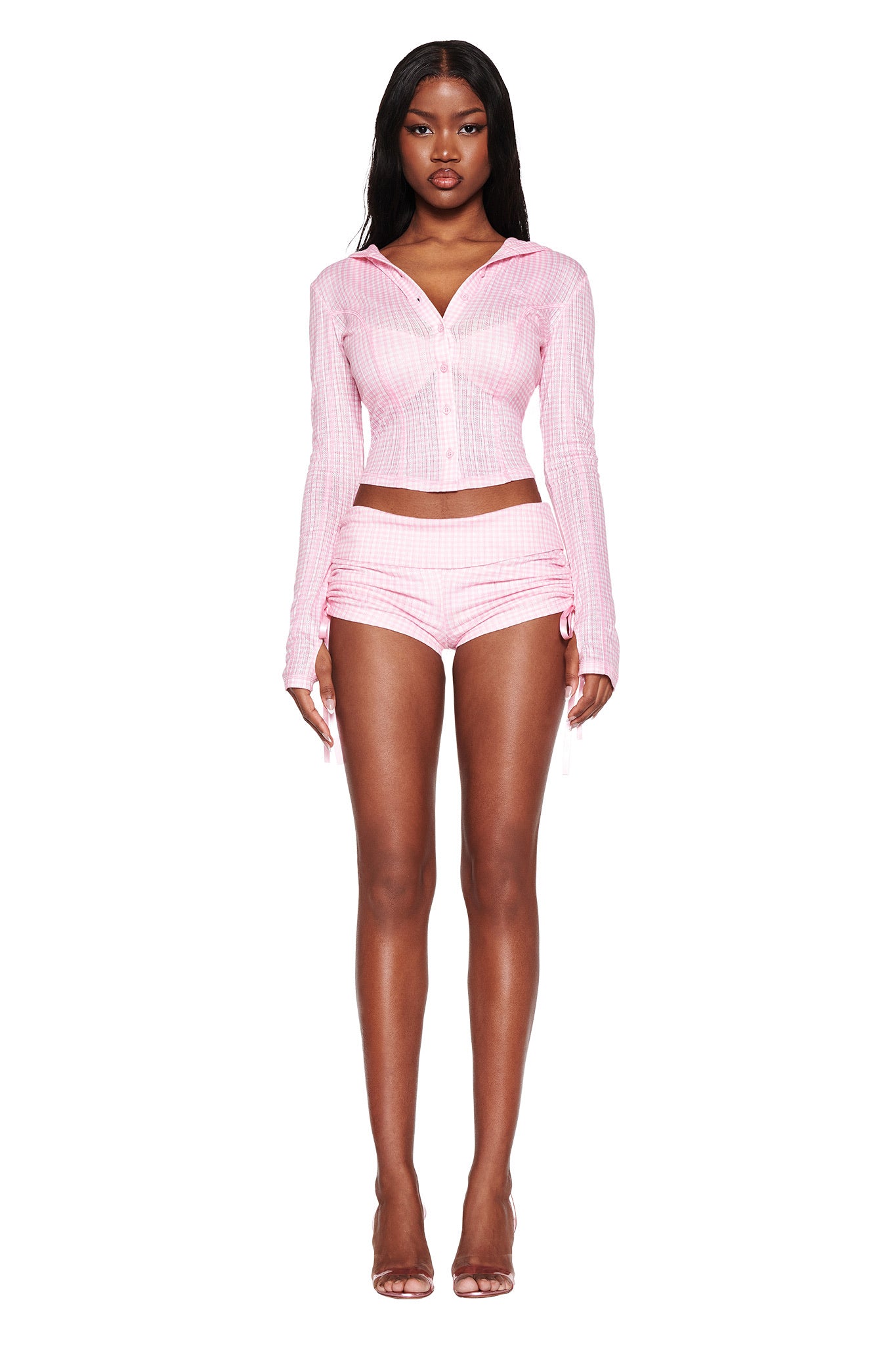 A model poses in the I.AM.GIA MIA SHORT - PINK, a baby pink gingham crop top with matching high-waisted shorts, featuring a checkered pattern and bodycon silhouette. She stands against a white background wearing clear high-heeled sandals.