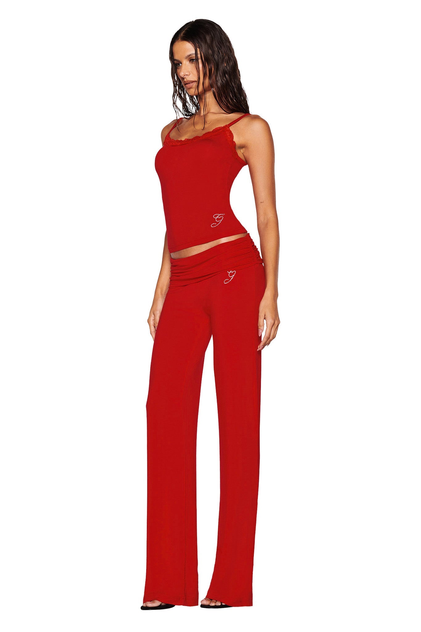 A woman is wearing the I.AM.GIA ERIN TOP in red, featuring a camisole silhouette with red lace trim and matching pants, posing against a white background. She has long, dark hair and stands with one hand on her hip. The outfit, crafted from bamboo fabrication, showcases small decorative elements near the waist.