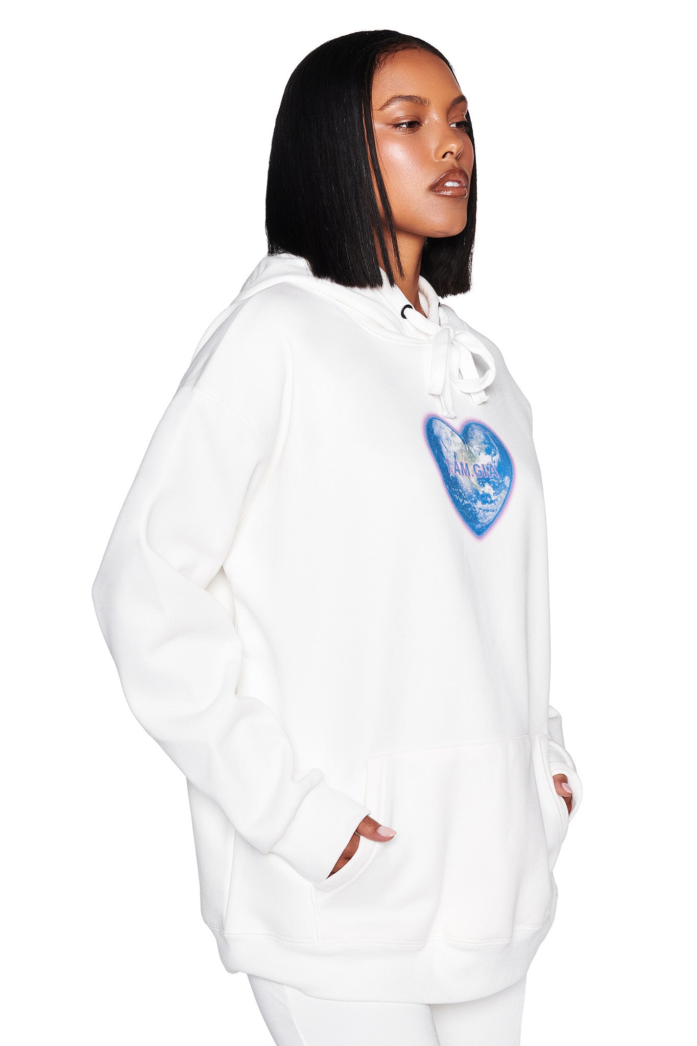 A person stands with hands in pockets against a white backdrop, wearing the I.AM.GIA Harmony Hoodie - White, featuring a blue heart graphic on the chest.