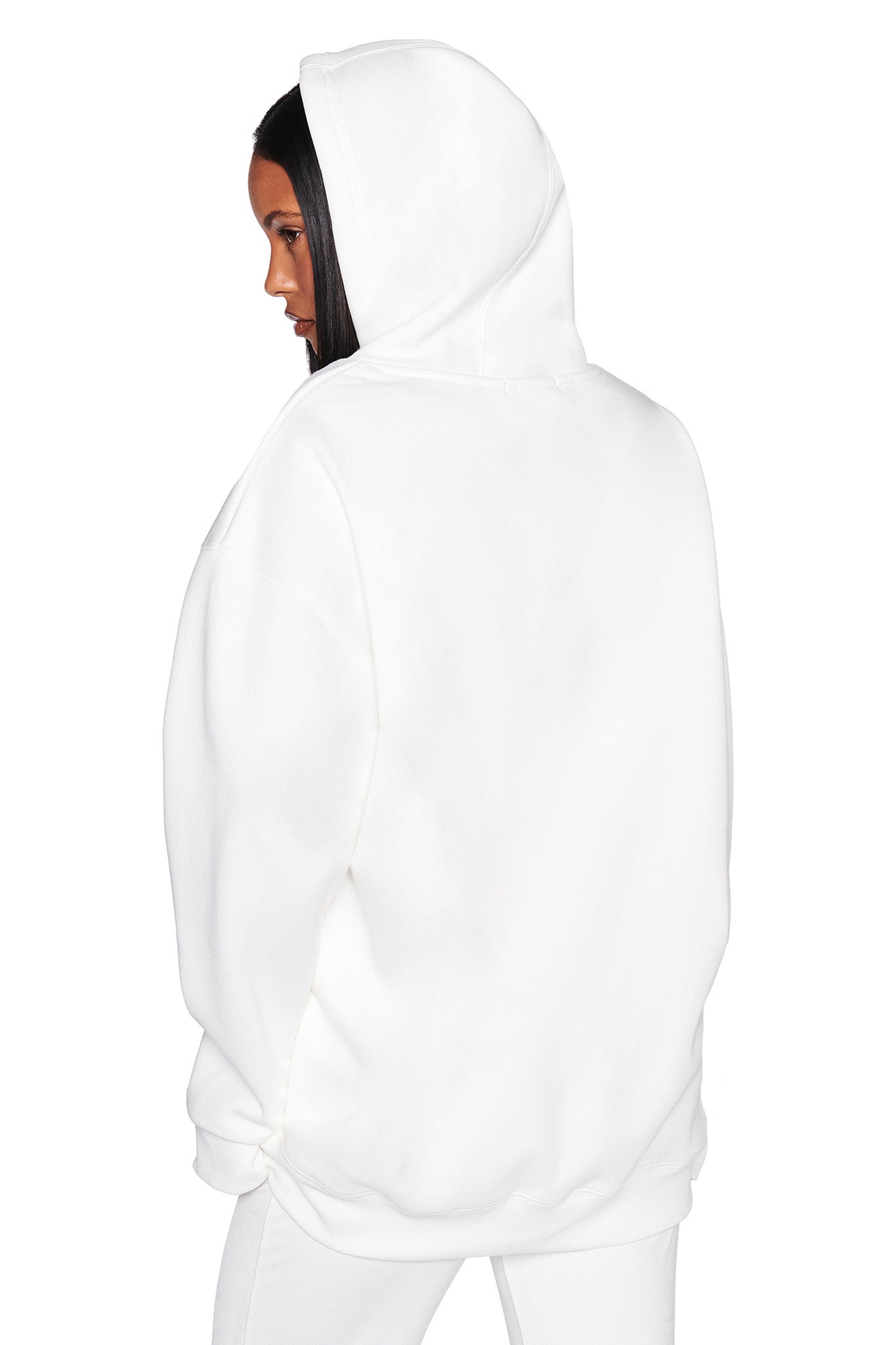 Someone with long hair wears an oversized I.AM.GIA HARMONY HOODIE - WHITE, hood up and facing slightly to the side against a plain white background, emphasizing the minimalist style of this chic sweatshirt.
