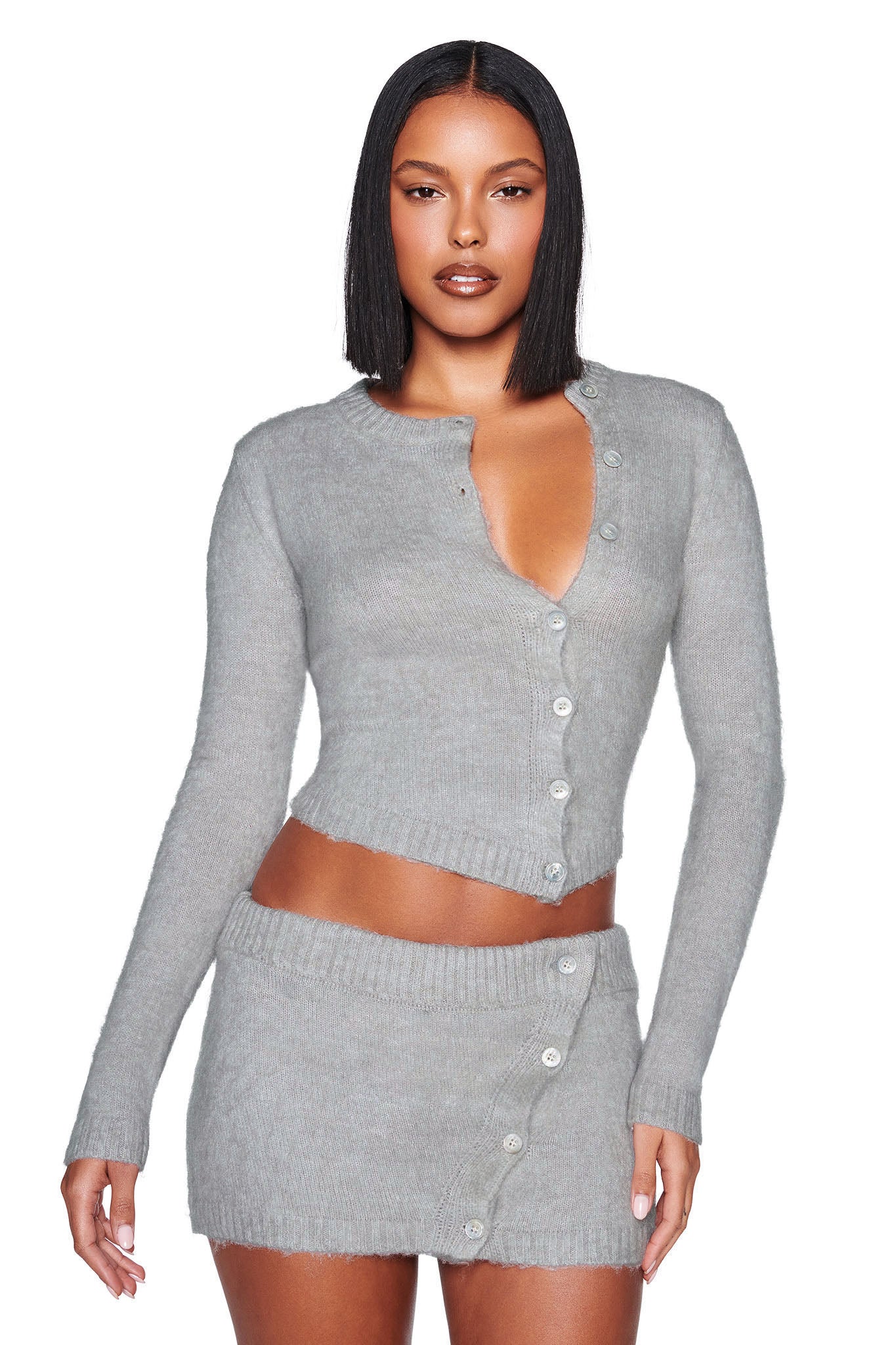 A woman has straight black hair, wearing I.AM.GIA's BEATRIZ SWEATER - GREY: MARLE. The relaxed-fit knit cardigan is light gray with long sleeves and a middle button closure, paired with a matching mini skirt featuring an asymmetrical hem and side buttons. Both have a ribbed texture.