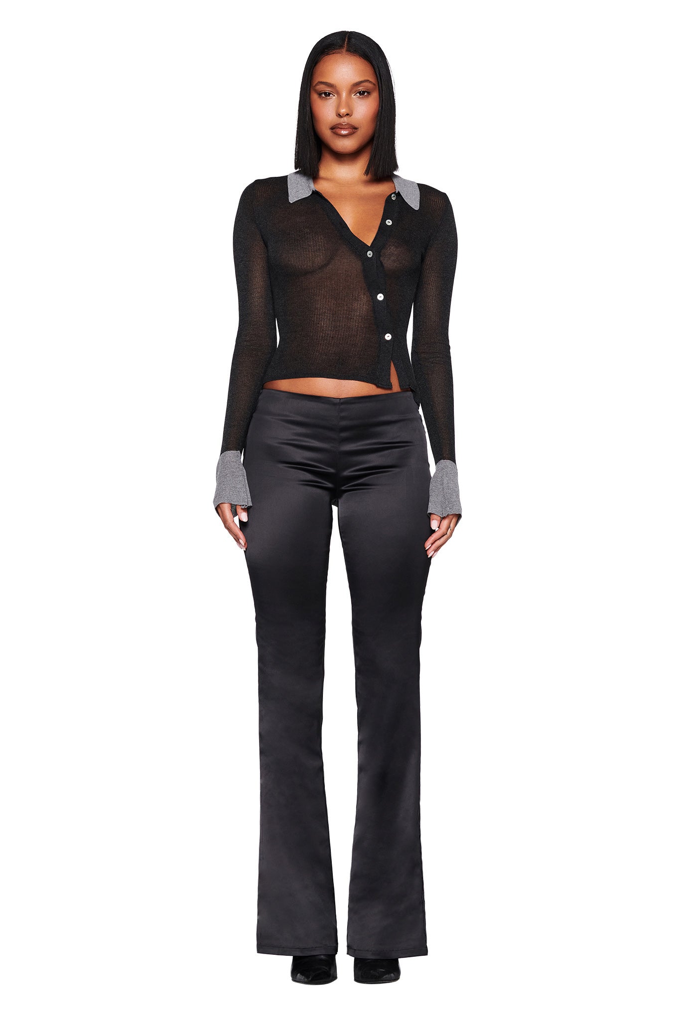 A person in an I.AM.GIA DAVINA TOP - BLACK, a fitted button-up sweater with pearl shell buttons and true knit fabrication, paired with black satin flared pants and black shoes, stands against a white background.