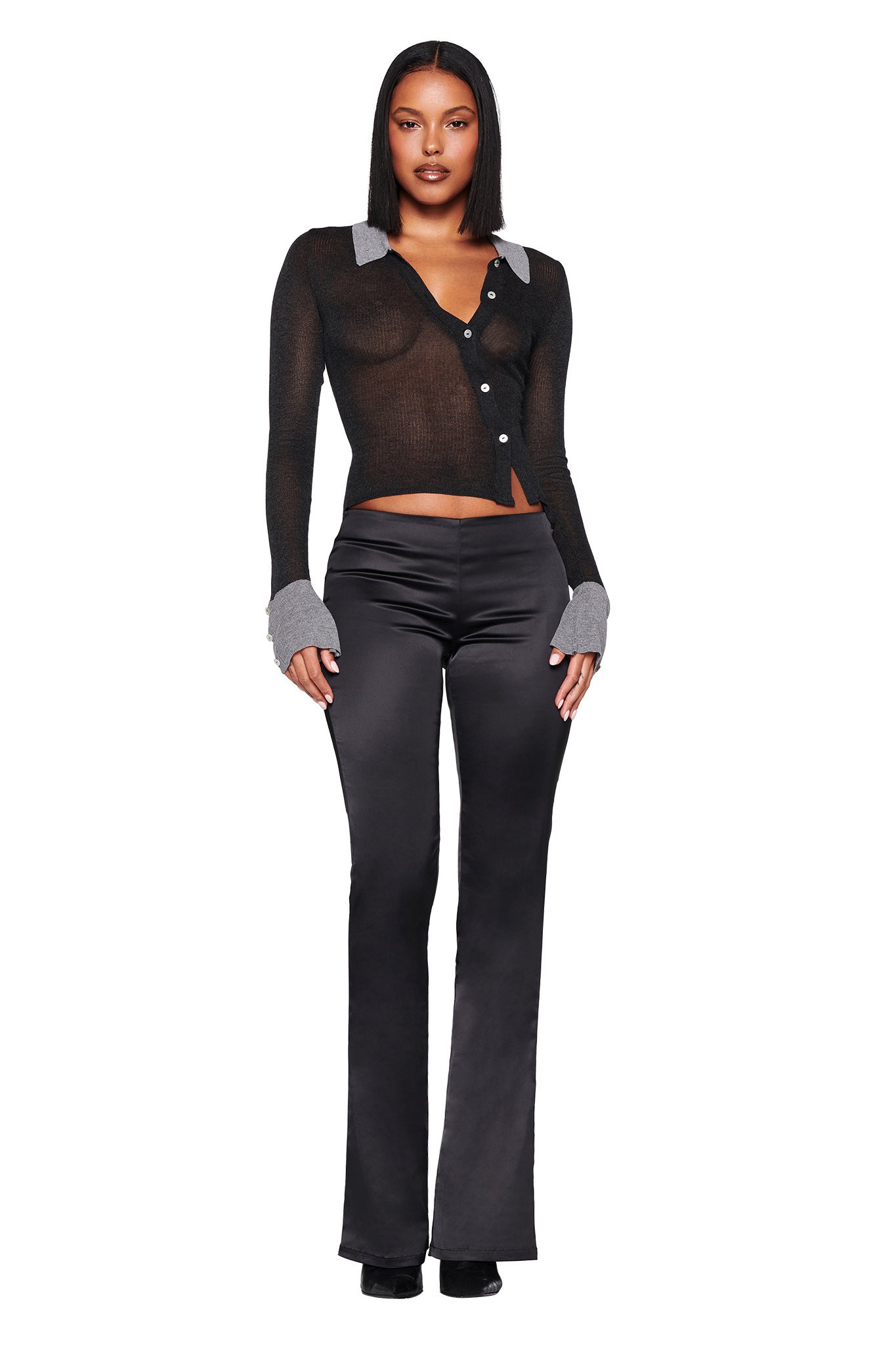 Wearing the chic I.AM.GIA Davina Top in black, with pearl shell buttons and grey cuffs and collar, they pair it effortlessly with satin black trousers. Their long, straight hair flows elegantly as they stand against a plain white background.