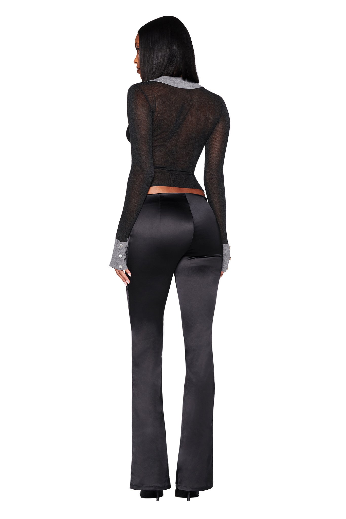A woman stands with her back turned, stylishly wearing the sheer black I.AM.GIA DAVINA TOP featuring pearl shell buttons, complemented by dark satin pants. Her straight hair flows to her shoulders against a simple white backdrop.