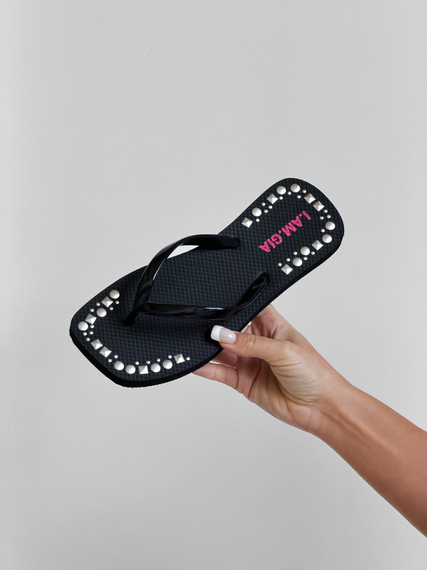 A hand holds the I.AM.GIA INKA STUDDED FLIP FLOP - BLACK, featuring pearl-like studs on the edge and "JAMAICA" in pink on its extra-soft foam footbed, set against a plain light background.