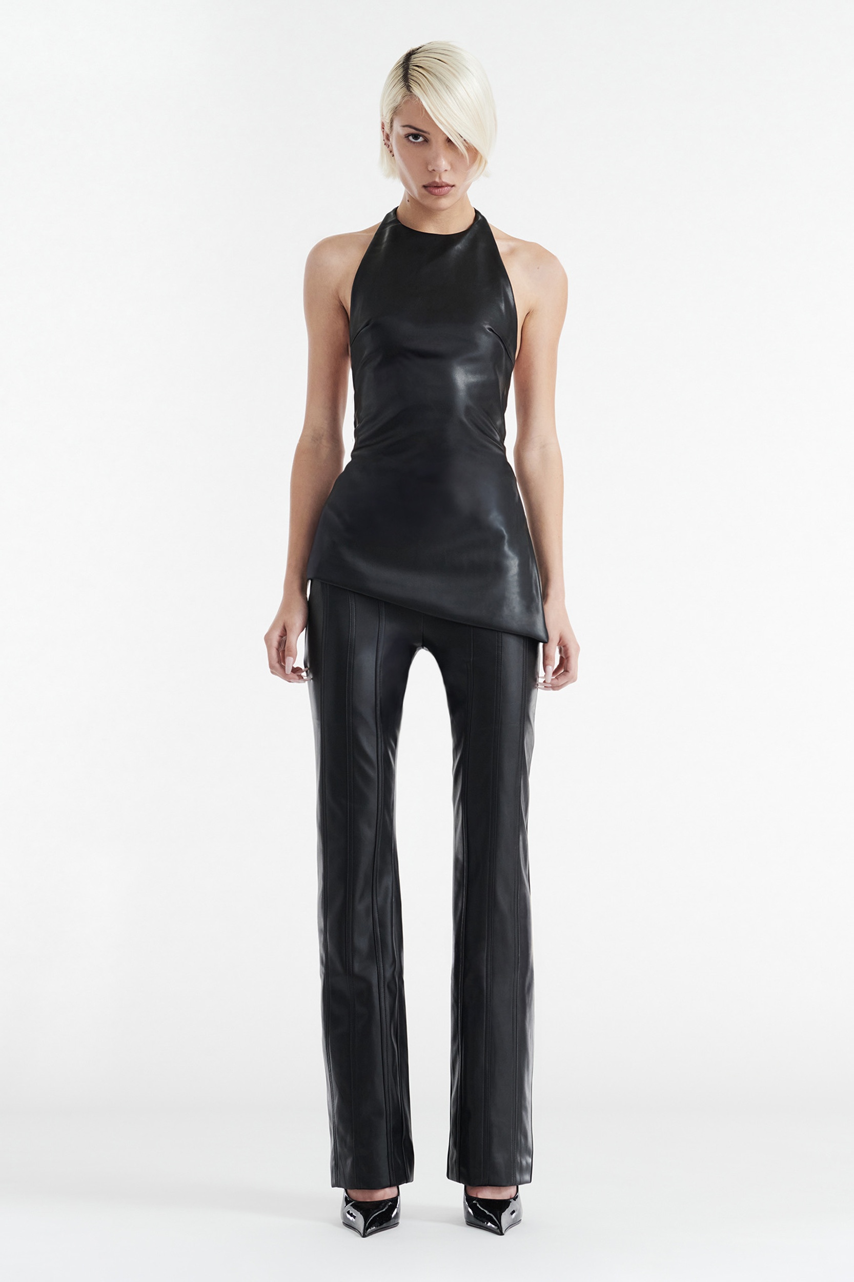 A person with short blonde hair stands against a white background, wearing the CDP INES Asymmetrical Faux Leather Top in black, paired with matching black leather pants and black pointed-toe heels.