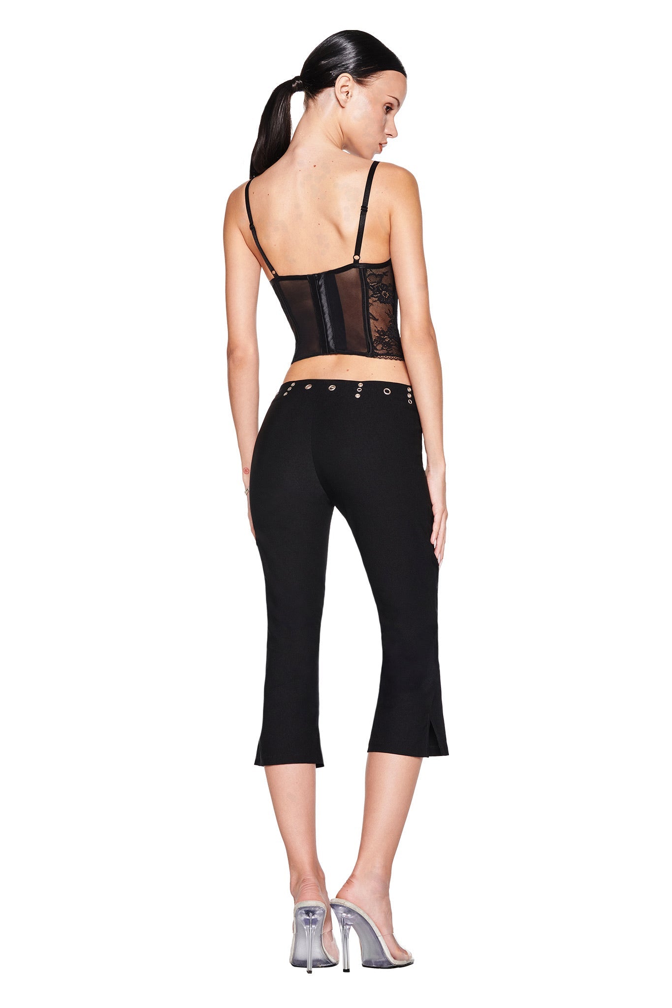 A woman with a ponytail faces away, wearing I.AM.GIA SHARAH CAPRI - BLACK pants with silver eyelet details and a low-rise waist, paired with a black lace corset top and clear heels, set against a white background.