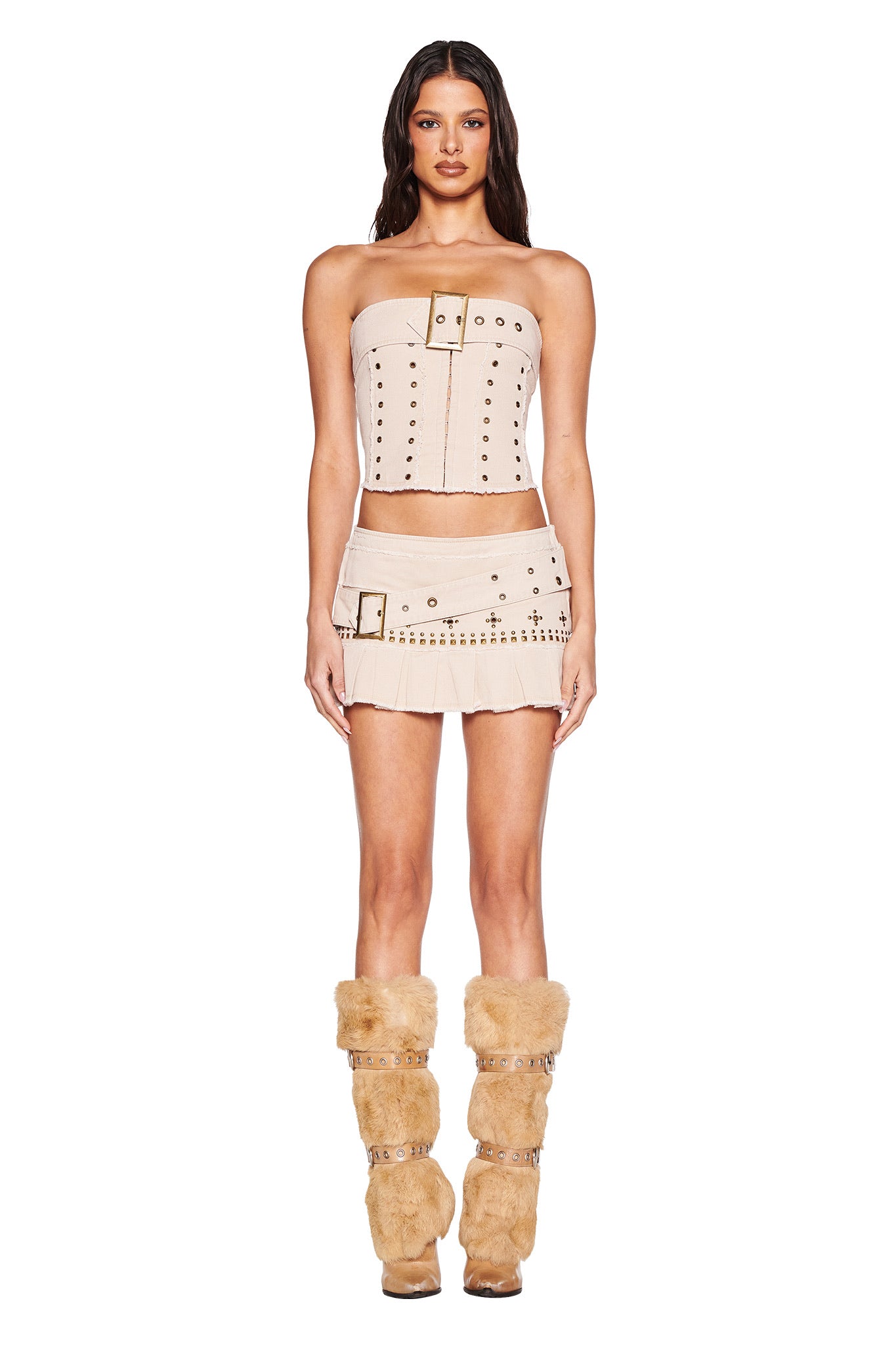 A woman faces forward against a plain white background, wearing the I.AM.GIA MASON CORSET in tan with matching mini skirt, both featuring metal details, and tan furry boots with buckled straps.