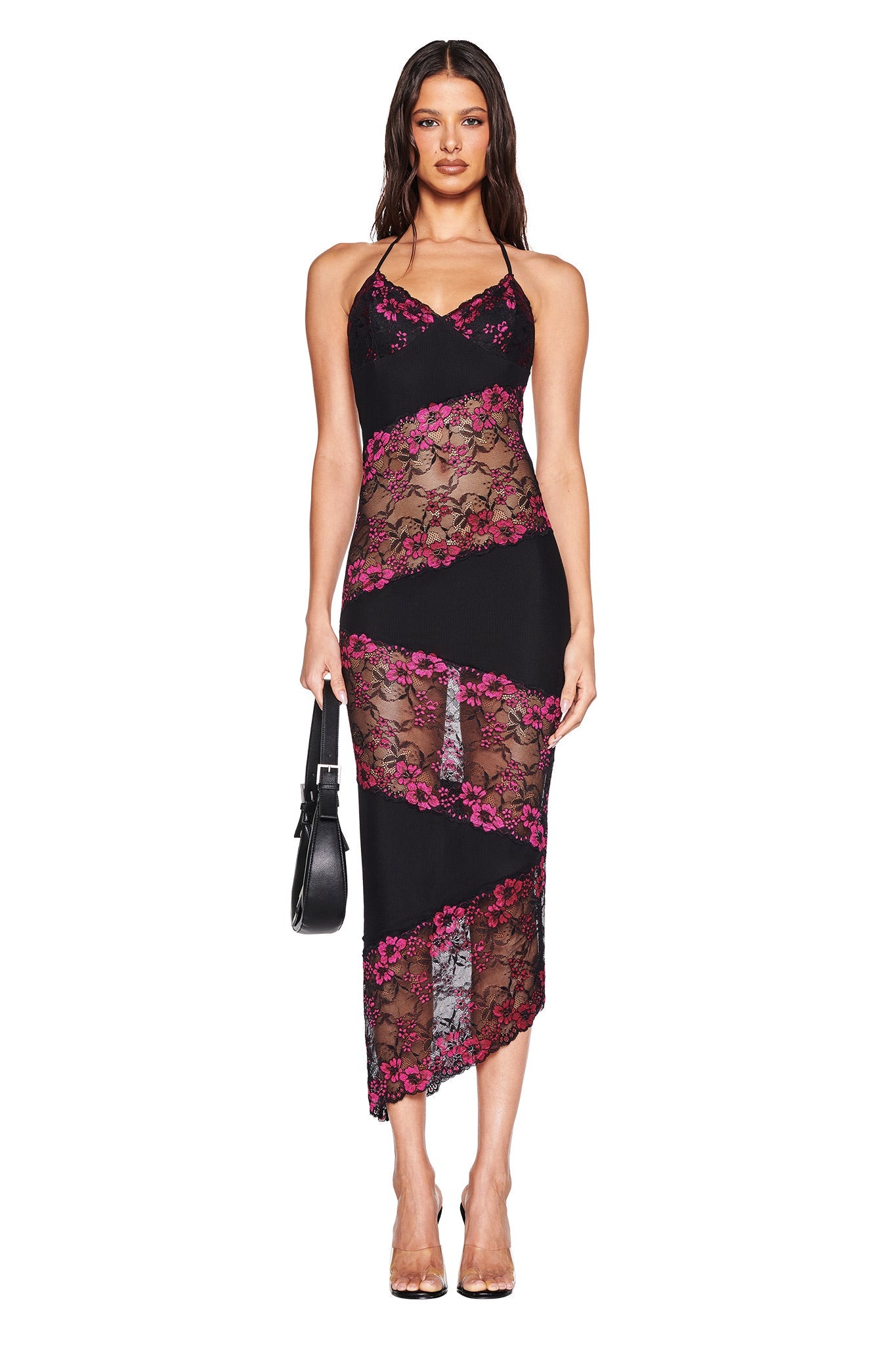 A woman faces forward in the I.AM.GIA PALOMA MIDI DRESS - BLACK, a sleeveless body-con style with sheer panels and an asymmetrical lace hem. She holds a black handbag, wears clear heels, and has long wavy hair.