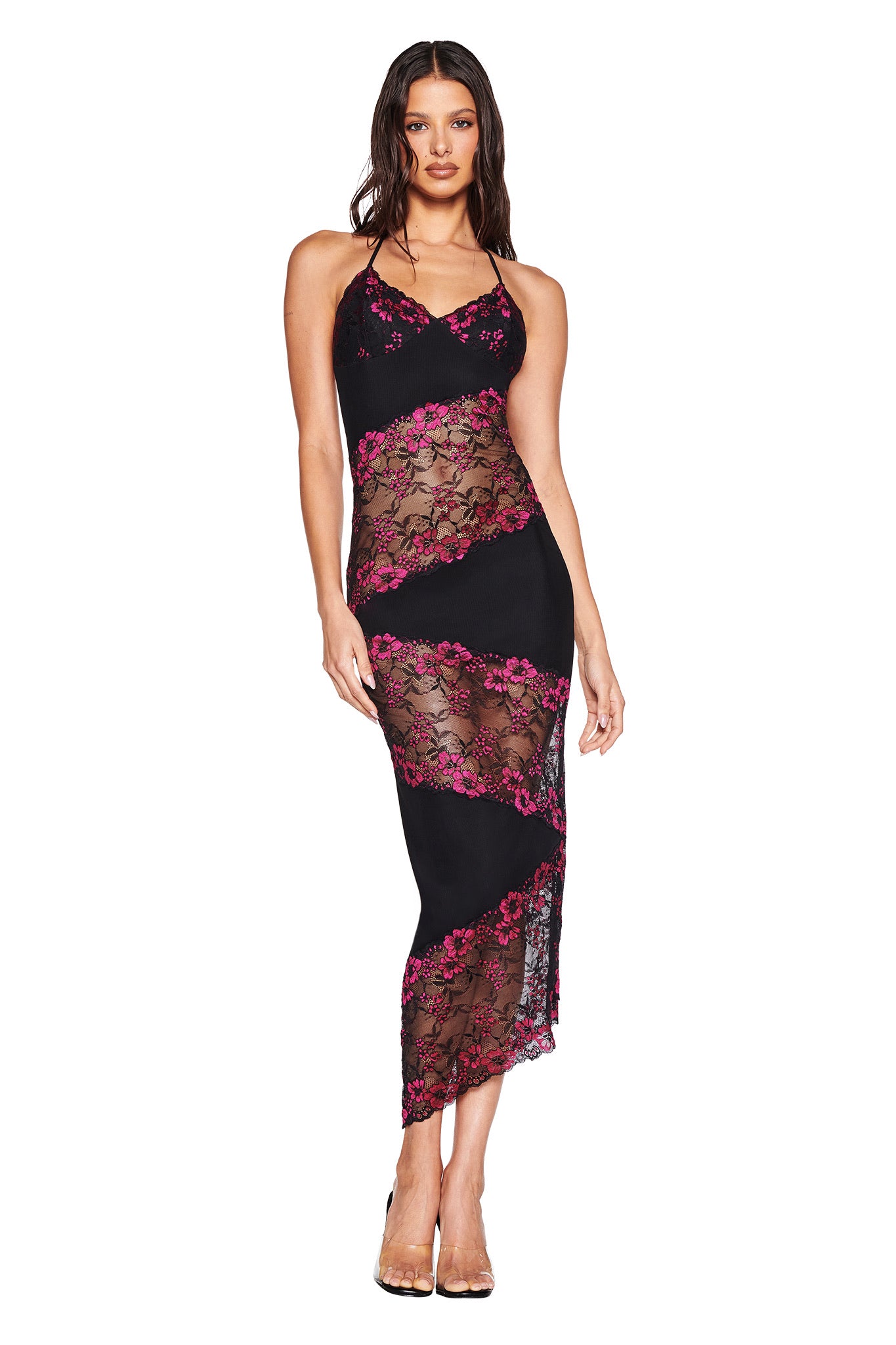 A woman wears the I.AM.GIA PALOMA MIDI DRESS in black, a body-con halter style with sheer diagonal lace panels, pink floral embroidery, an asymmetrical lace hem, and clear high heels against a white background.
