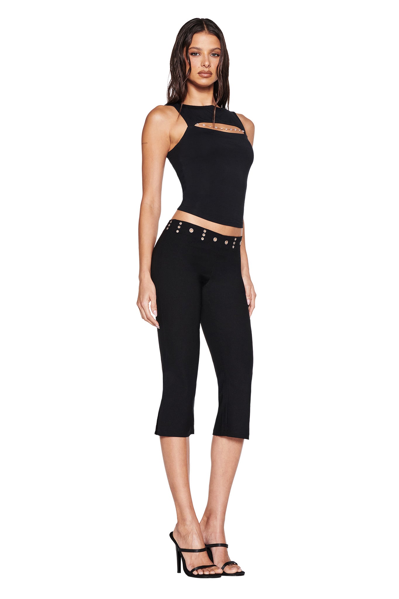 A woman wears the I.AM.GIA SHARAH CAPRI in black—sleek slim capri pants with button details, a low-rise waistband, and mid-stretch fabric—paired with a black sleeveless cutout top and high-heeled sandals against a white background.