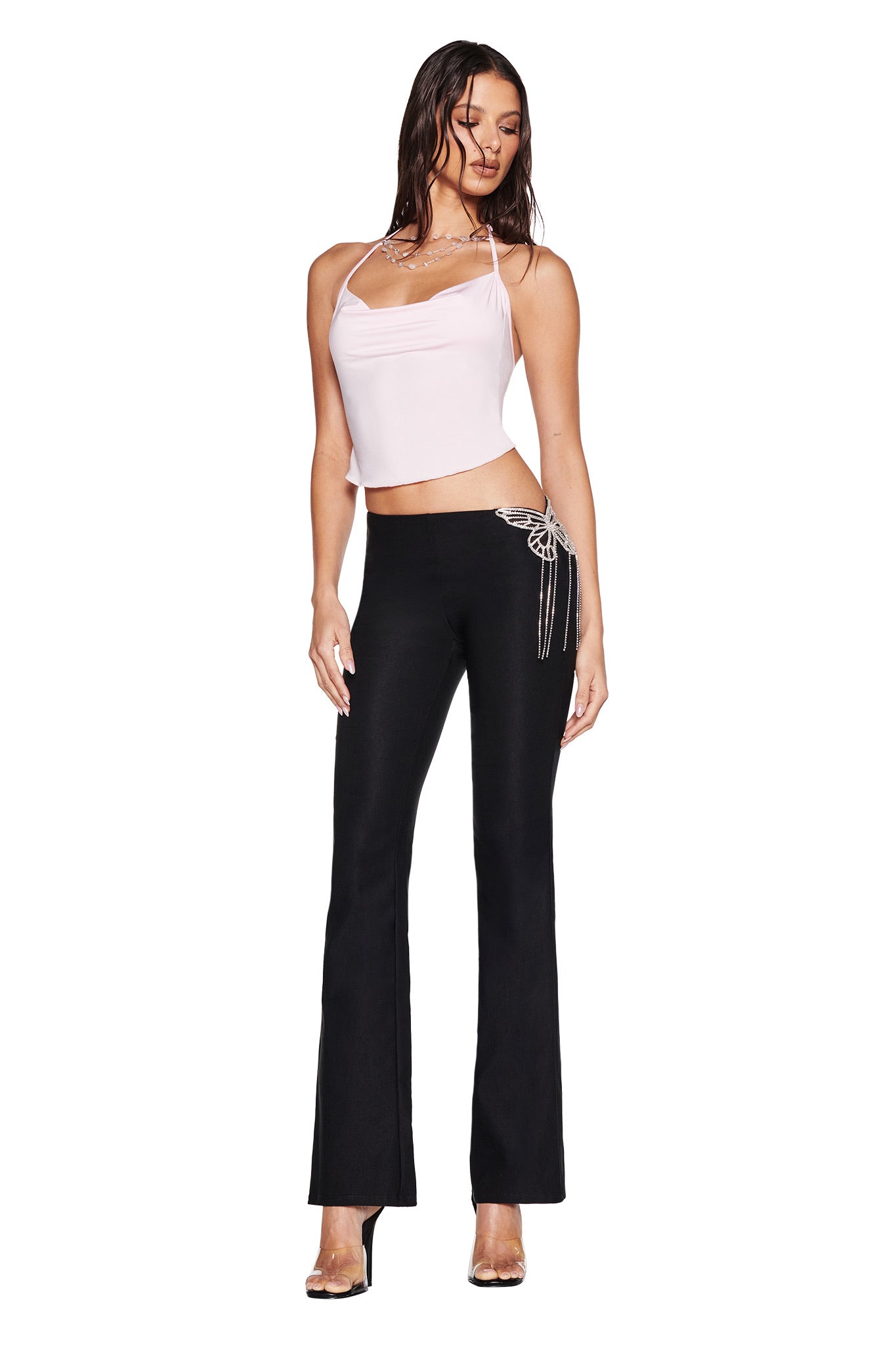 A woman wears the I.AM.GIA SORREL TOP in pink, a baby pink backless halter, styled with black flared pants featuring jeweled hip detail and high heels, posing against a white background.