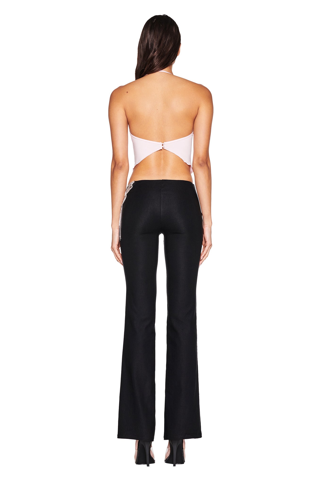 A woman faces away, wearing the I.AM.GIA SORREL TOP - PINK, a baby pink backless halter top with a crystal butterfly chain, paired with high-waisted black flared pants and heels against a plain white background.
