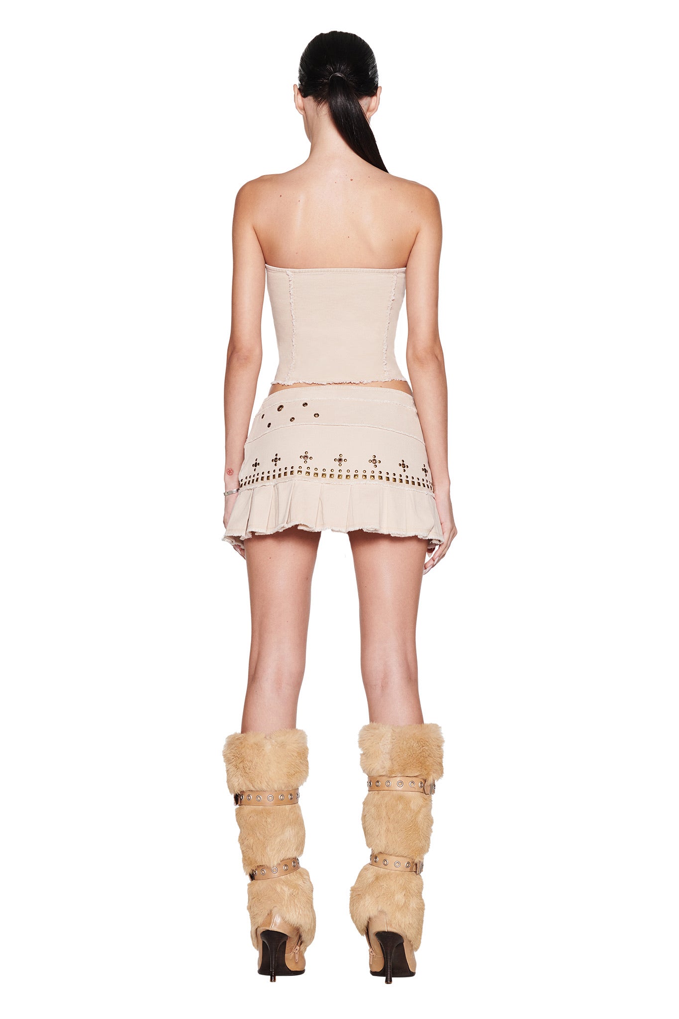 A woman with long dark hair in a ponytail faces away, wearing the I.AM.GIA MASON CORSET - TAN mini dress with pleated skirt and stud details, paired with tall tan furry heeled boots.