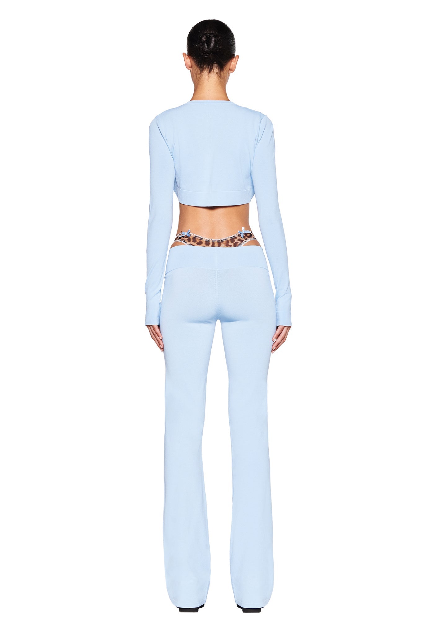 A woman stands facing away, wearing a long-sleeve, baby blue crop top and the I.AM.GIA AUDRINA PANT in powder blue. The high-stretch flare pants feature a patterned chain belt around the low-rise waist. Her hair is pulled back into a sleek updo as she poses against a plain white background.