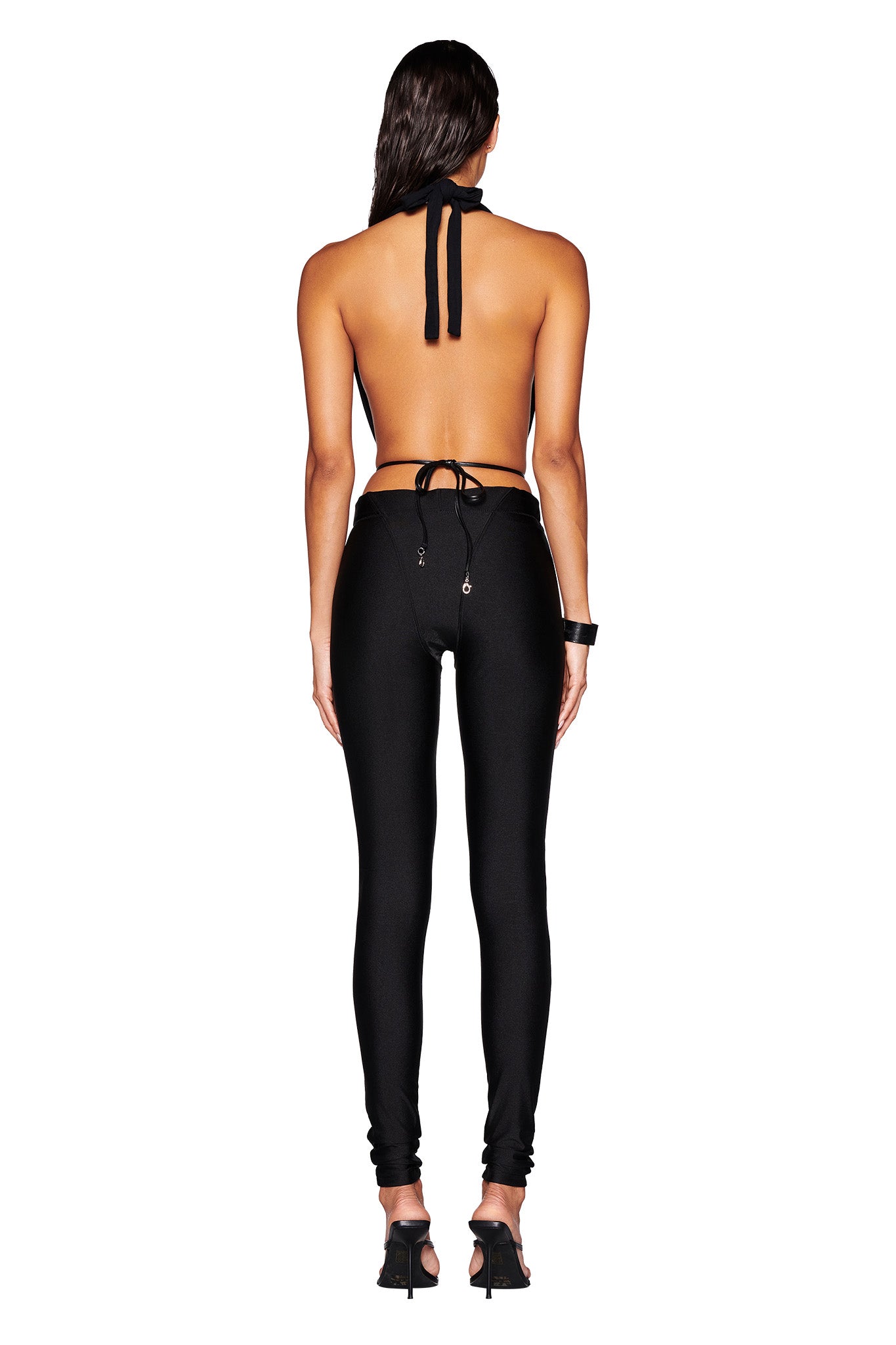 A person poses with their back to the camera, highlighting the MIKAYLA TOP in black by I.AM.GIA, paired with fitted black pants. They complete the look with black high-heeled shoes and a bracelet on their right wrist, all set against a crisp white background.