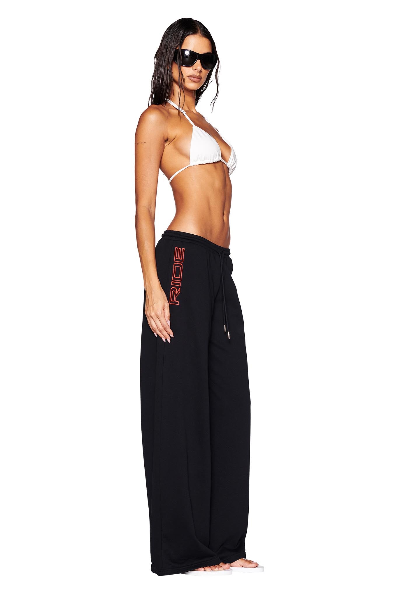 A woman confidently stands against a white background wearing the JUSTINE PANT in black by I.AM.GIA, which features the word "RIDE" in bold red letters. Her outfit is completed with a chic white bikini top and striking black sunglasses that highlight her wet, flowing hair. She embraces barefoot style with a low-rise waist fit.