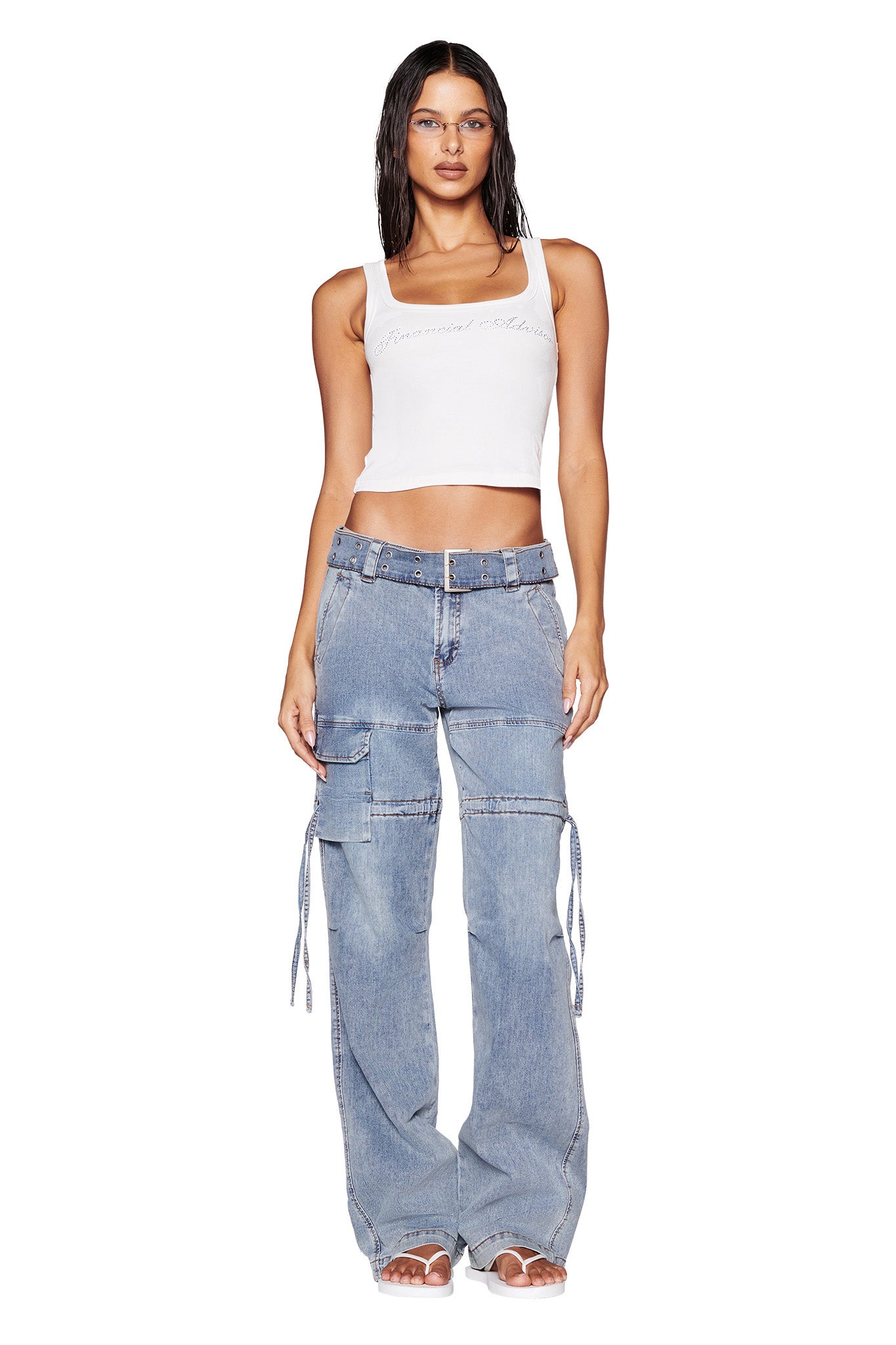 A woman stands facing the camera, wearing a white sleeveless crop top and light blue NAIMA CARGO PANT - DENIM by I.AM.GIA, which feature multiple pockets and straps. The relaxed silhouette complements her long, dark, wet-look hair. She completes the look with white open-toe shoes, all set against a plain white background.