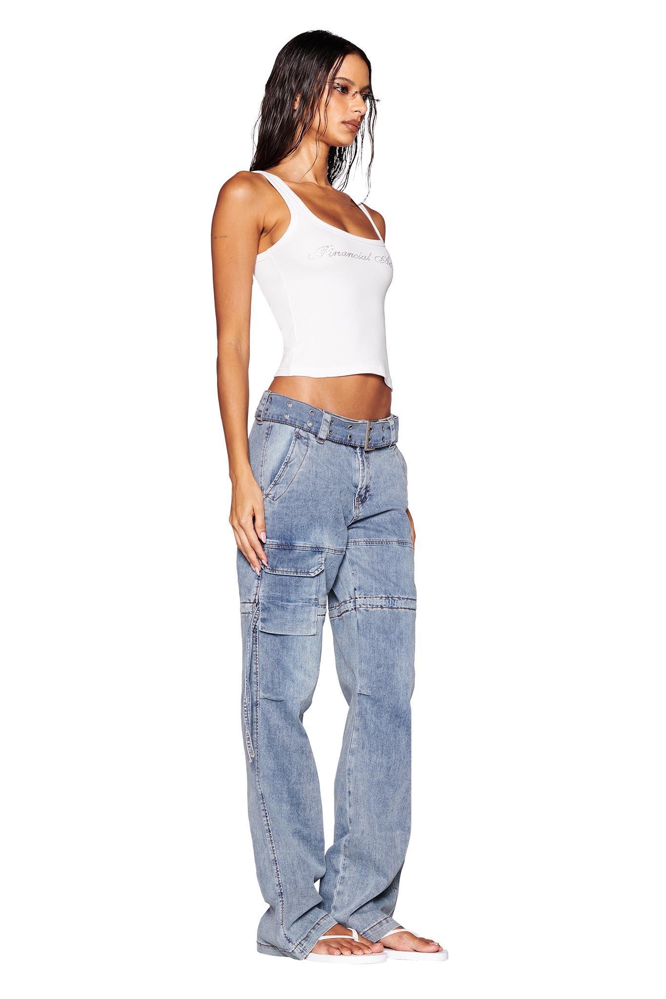 A woman with long hair stands against a white background, wearing a white crop top and light blue NAIMA CARGO PANT - DENIM by I.AM.GIA, which feature pockets and a low-rise waist. With one hand by her side, she looks to the left while showcasing the relaxed silhouette of her outfit enhanced by white flip-flops.