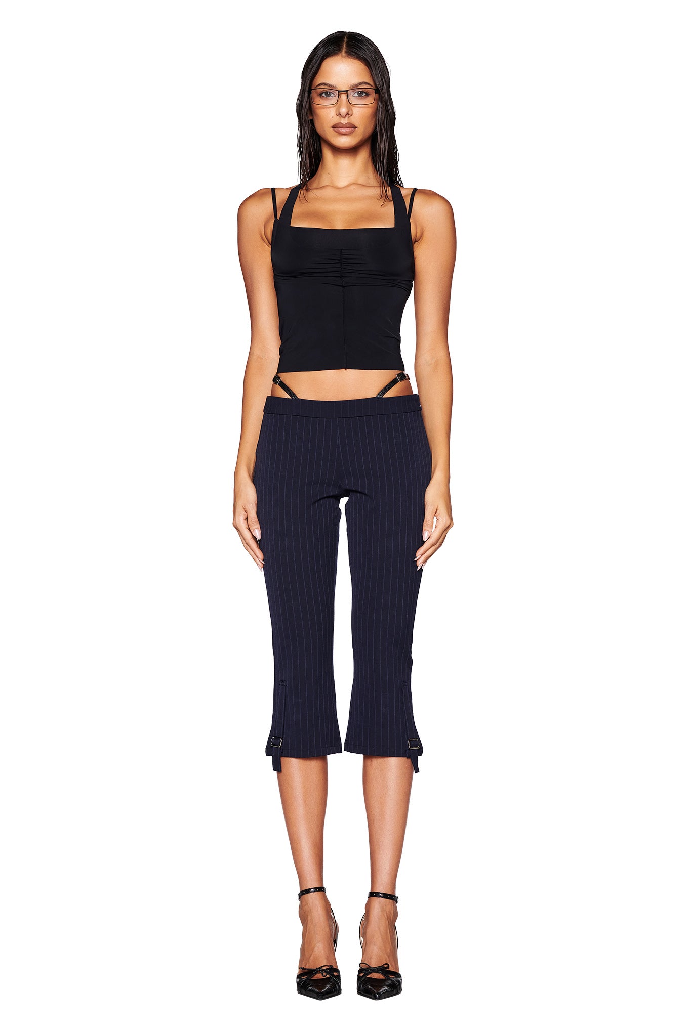 A woman with long hair is wearing a fitted black top paired with the MARRION CAPRI - BLUE: PINSTRIPE pants from I.AM.GIA, featuring a low-rise waist. She stands against a plain white background, looking directly at the camera with her arms at her sides, and sports black high heels adorned with faux leather straps.