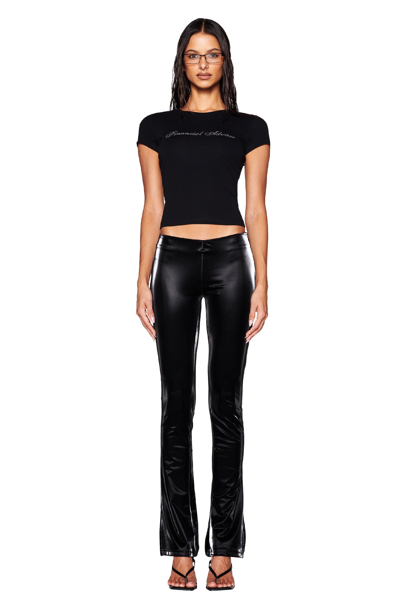 A person with long hair stands against a white background, wearing glasses, a black fitted t-shirt with text, the I.AM.GIA HARLO PANT in black featuring a shiny faux leather finish with flared legs and a low-rise waist, and black strappy sandals.