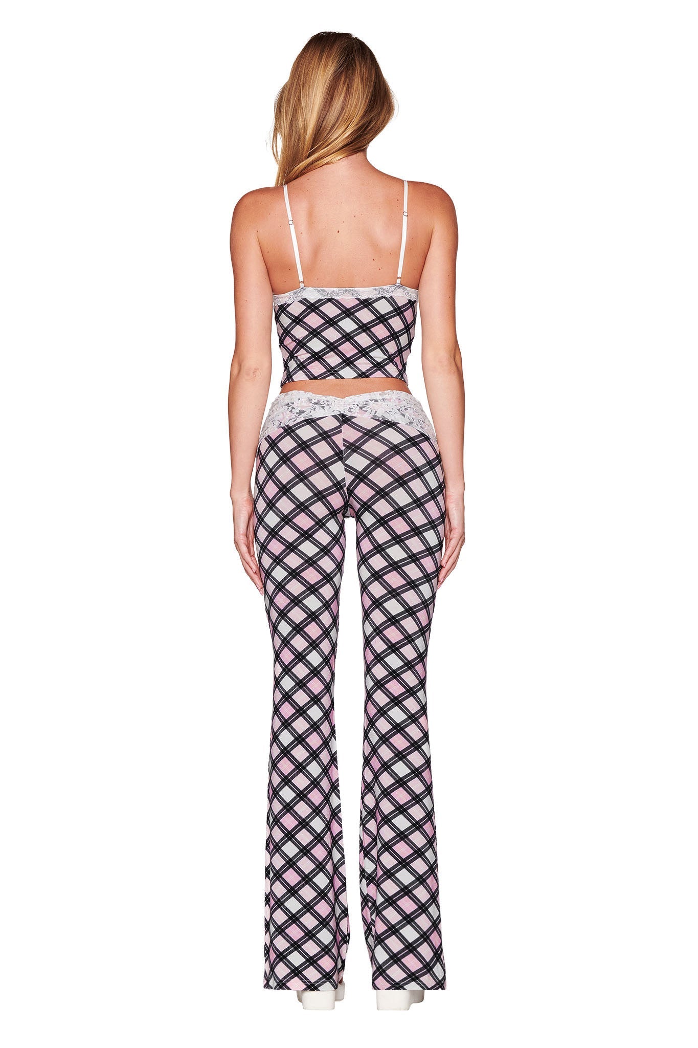 A person with long hair stands facing away, showcasing the KENDRA PANT - PINK: PLAID from I.AM.GIA in a fashionable low-rise flared silhouette. The ensemble, featuring black, white, and pink hues, is elegantly paired with white platform heels for a stylish finish.